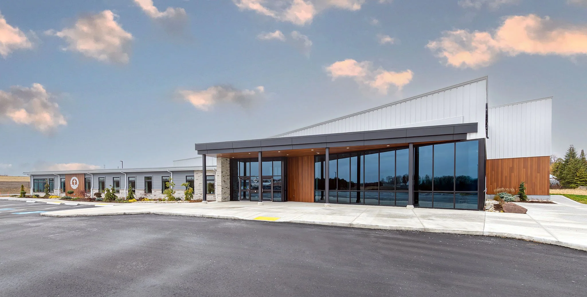 Modern commercial building with large glass windows, wood and stone accents, surrounded by a parking lot and landscaped area, under a partly cloudy sky.