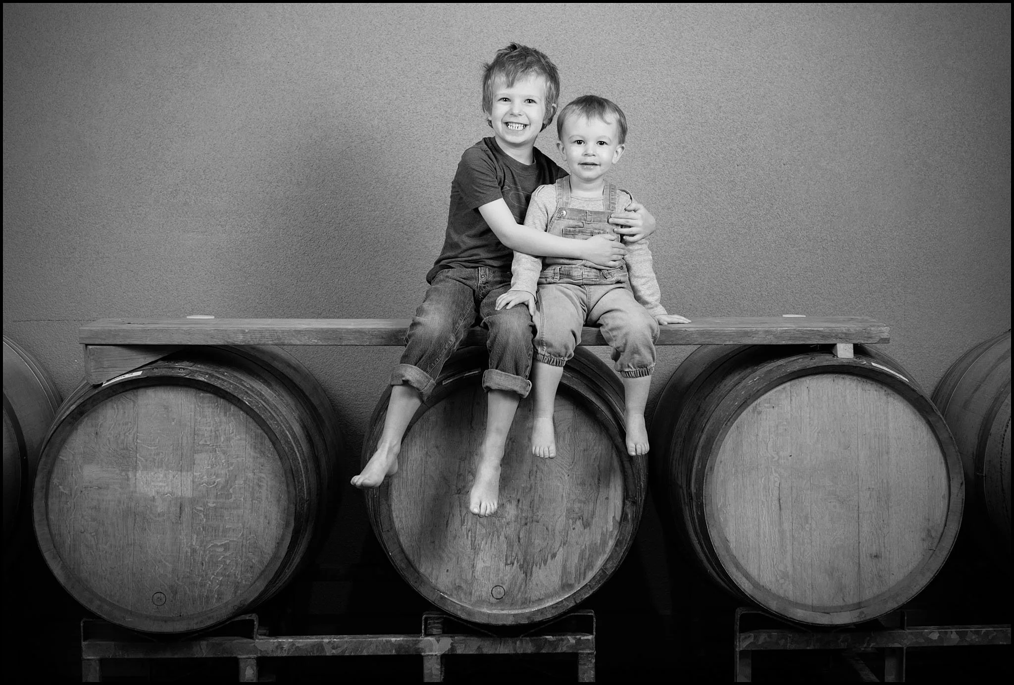 Two young boys sitting on large wooden barrels, one embracing the other, in front of a textured wall.