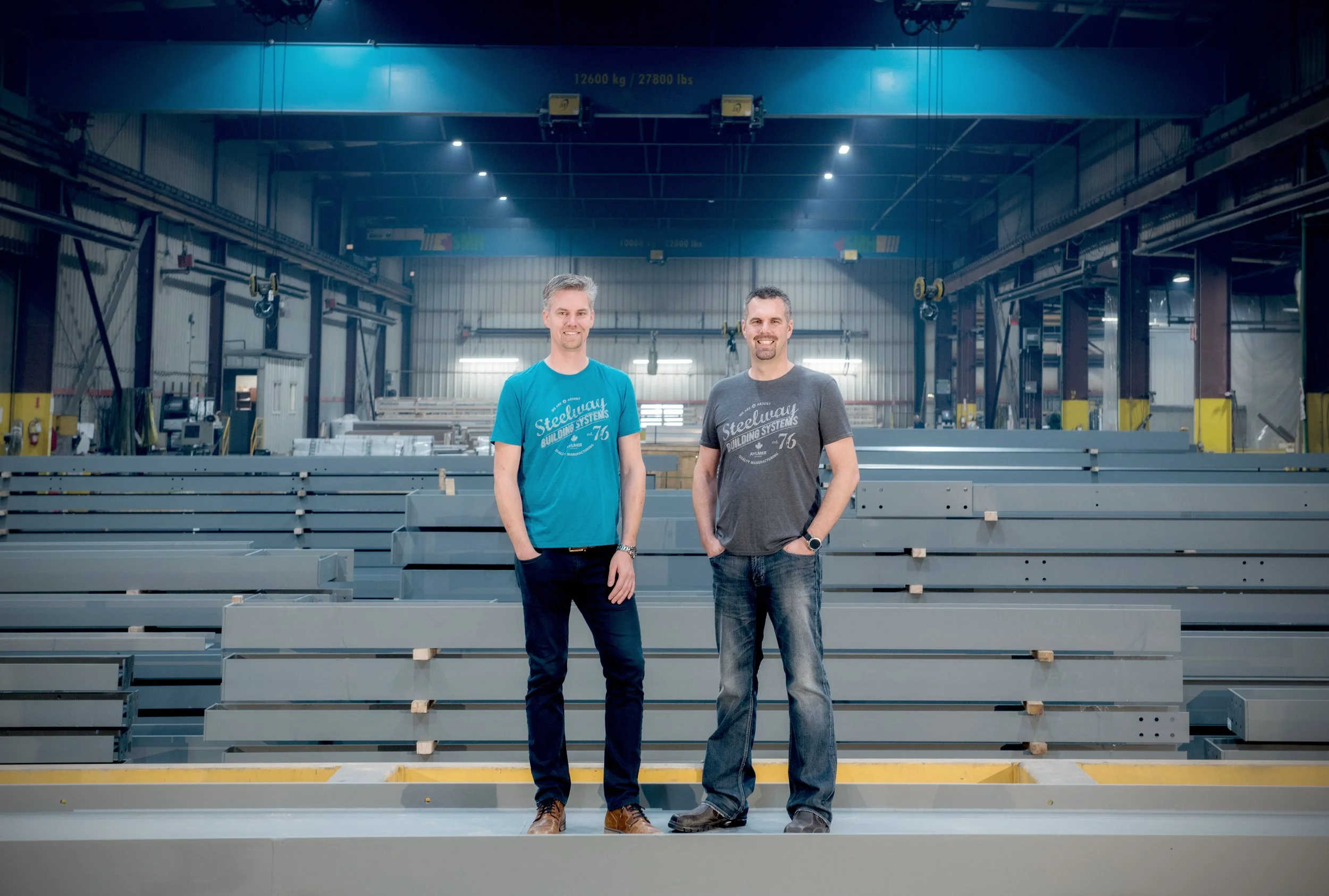 Two men standing in an industrial warehouse or manufacturing facility, smiling at the camera, surrounded by large metal construction elements, with a crane overhead.