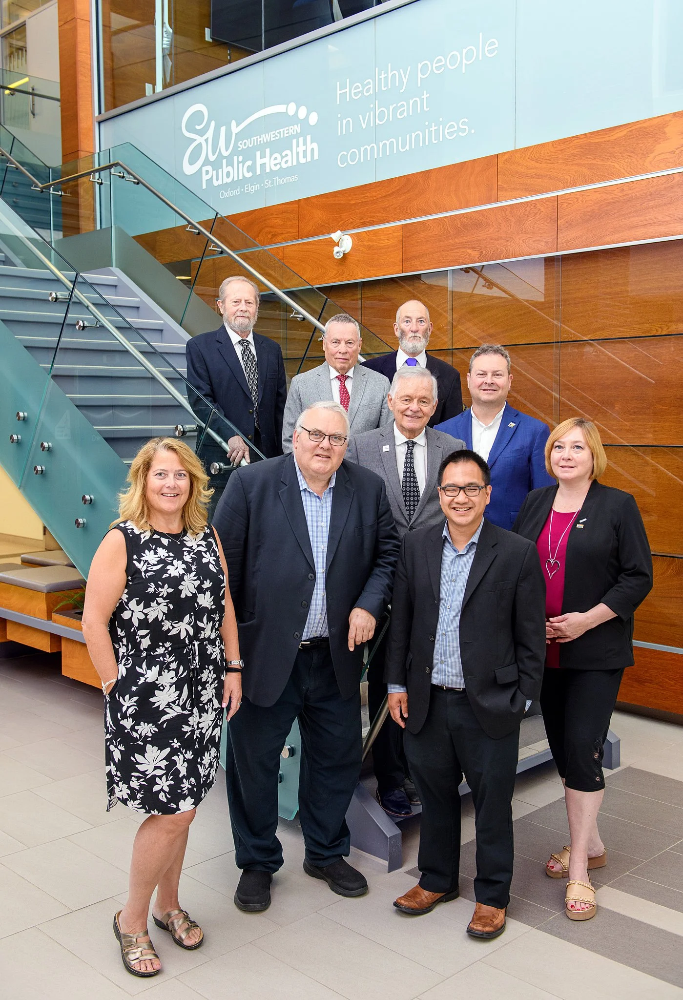 Group of nine diverse professionals standing in front of a staircase inside a building with wood-paneled walls. They are dressed in business and business casual attire, smiling at the camera. A large sign behind them reads "Southwestern Public Health
