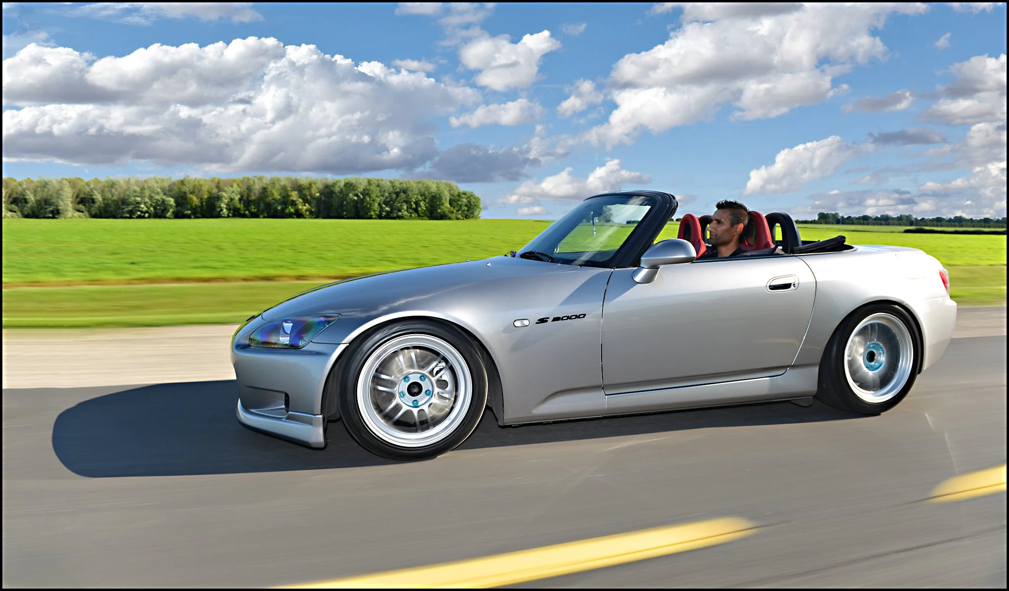 A silver convertible sports car driving on a road with a male driver and red and black seats, green fields and trees in the background, under a partly cloudy sky.