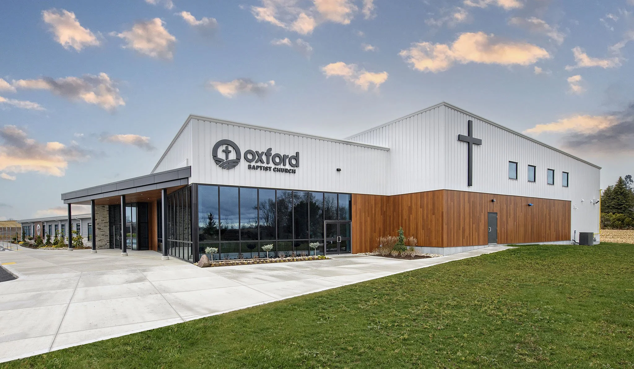Modern church building with sign reading "Oxford Baptist Church," featuring a cross symbol, large glass windows, and a landscaped exterior, under a partly cloudy sky.
