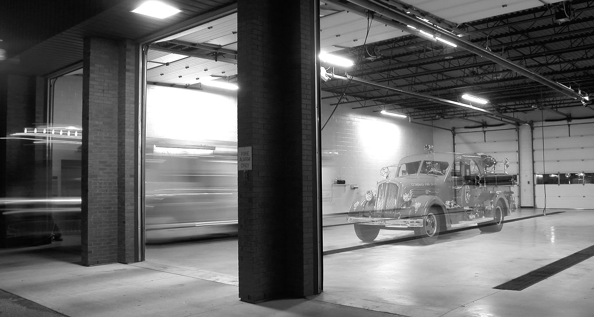 Black and white photo of a fire station or garage with a vintage fire truck inside, seen through large glass windows.