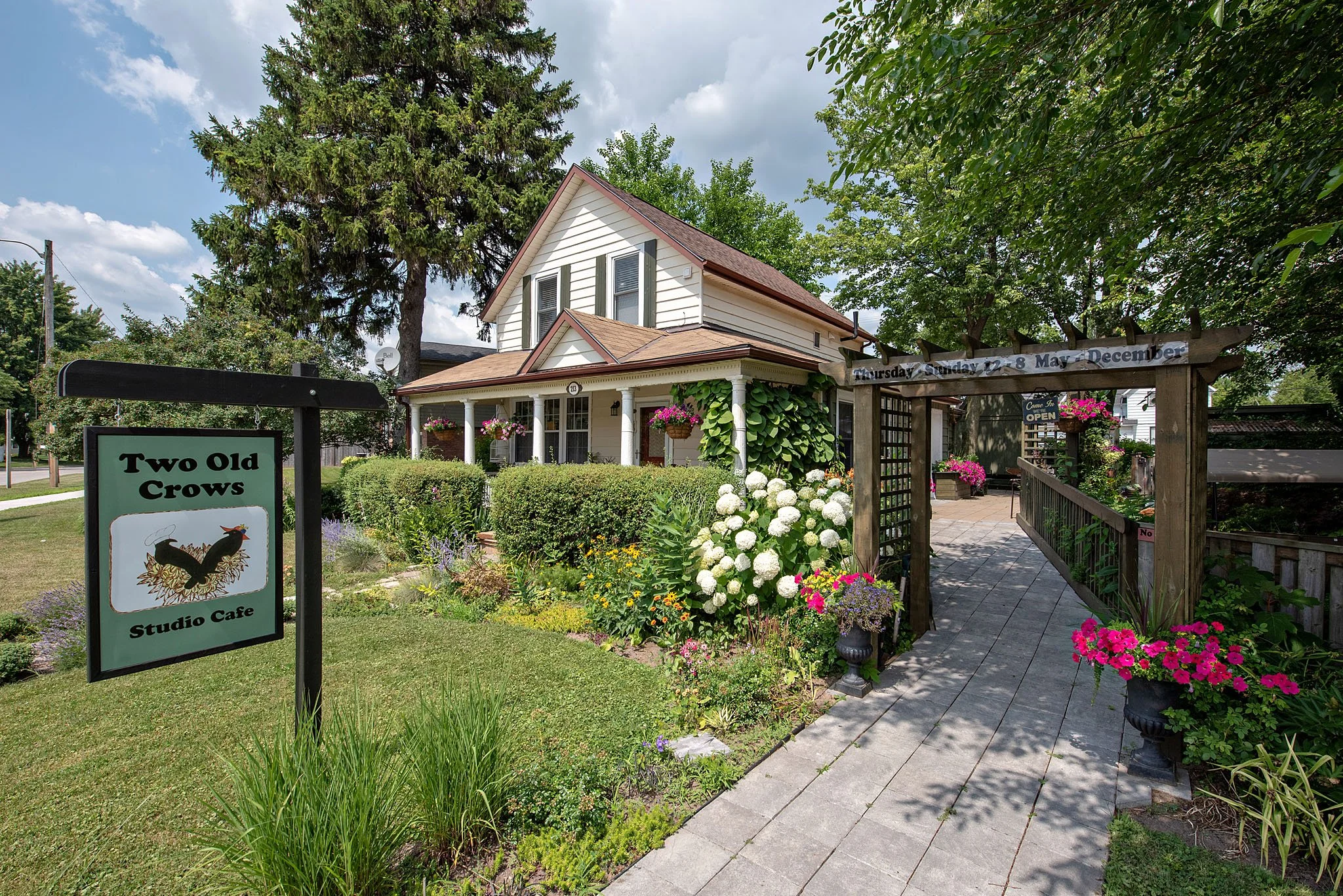 A charming white house with a porch, surrounded by colorful flowers and greenery, with a garden and a sign that reads 'Two Old Crows Studio Cafe' and neighboring signs indicating hours and open status.