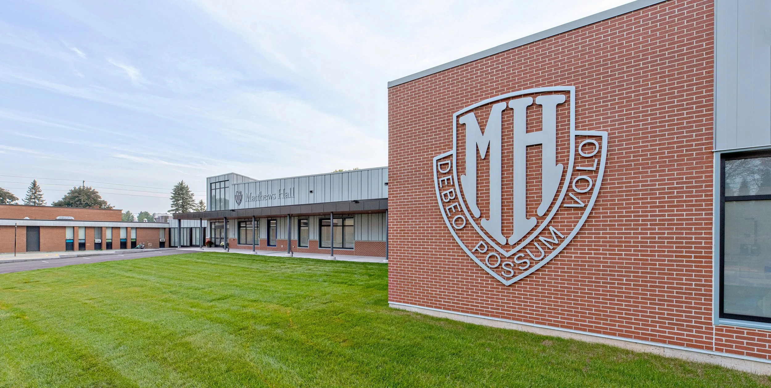 Exterior of a school building with a large emblem reading 'MH' and the words 'DeBeno Post' on a brick wall, and 'Madrowns Hall' on a section of the building, with a grassy lawn in the foreground.