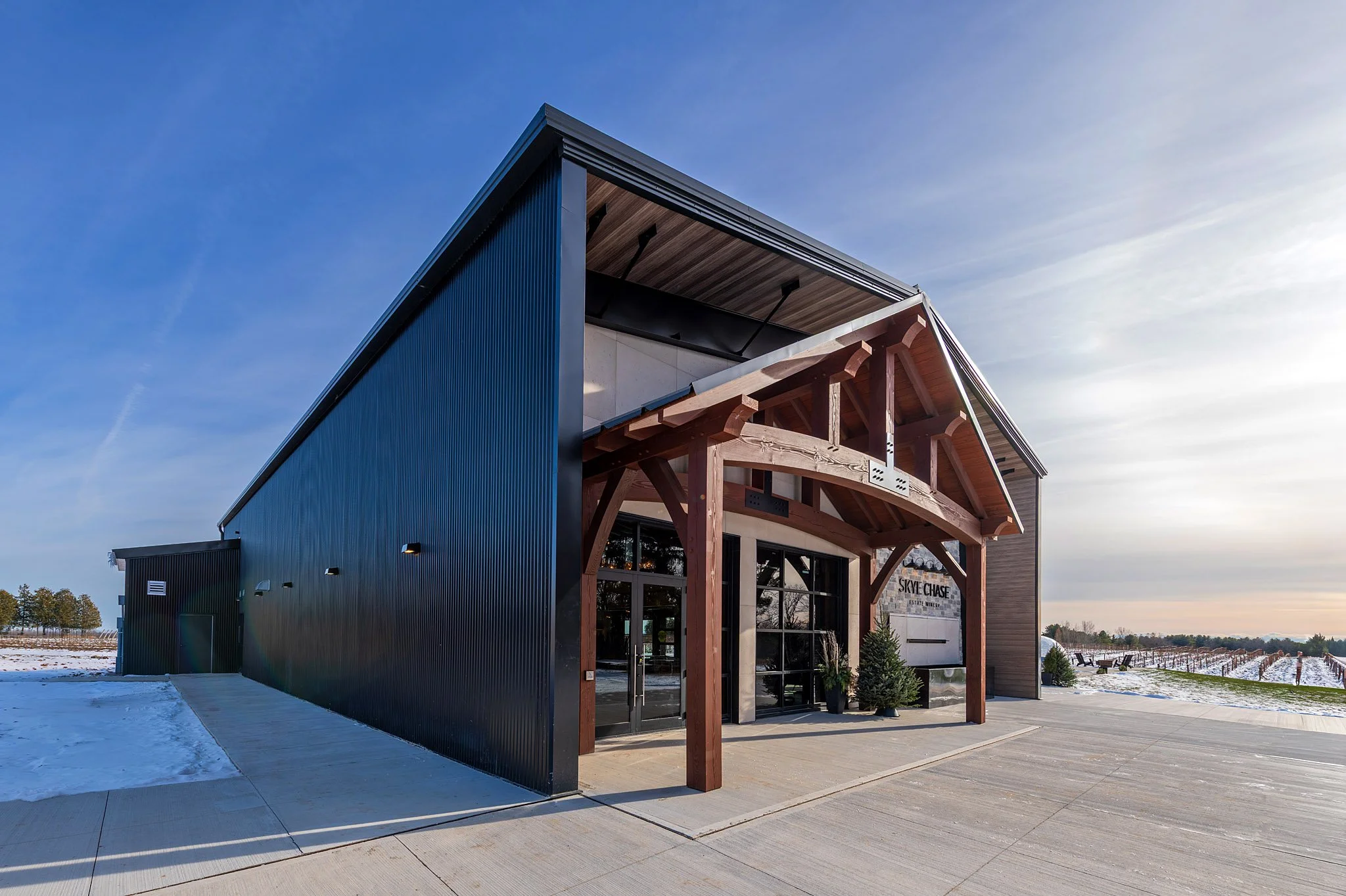 A modern building with a mix of blue metal siding and natural wood accents. The building has large glass doors and windows and is situated outdoors on a concrete patio. There is snow on the ground and a clear blue sky overhead.