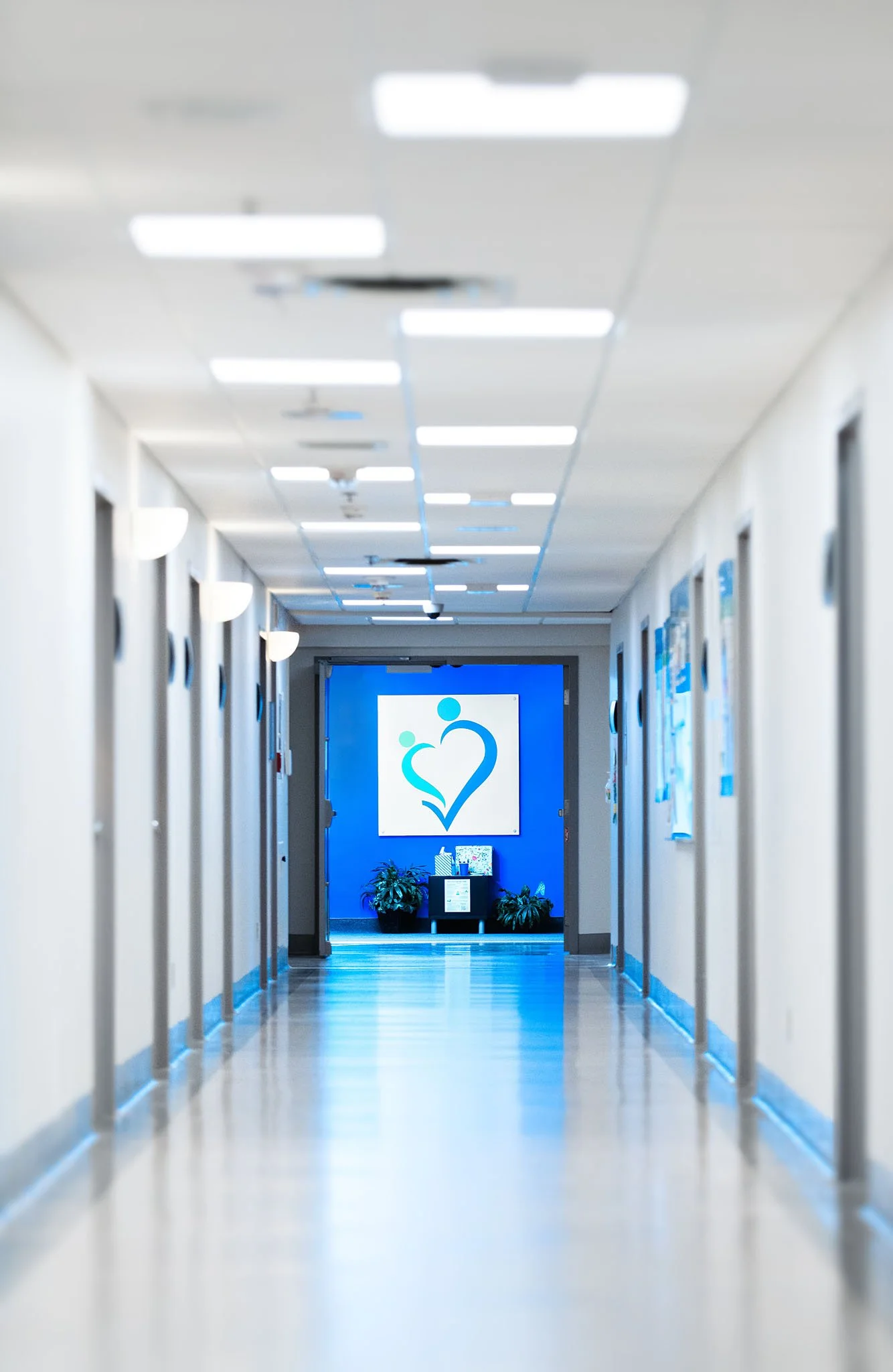 Long, empty hospital corridor with a blue wall at the end, decorated with a white logo of a heart with two smaller hearts inside.