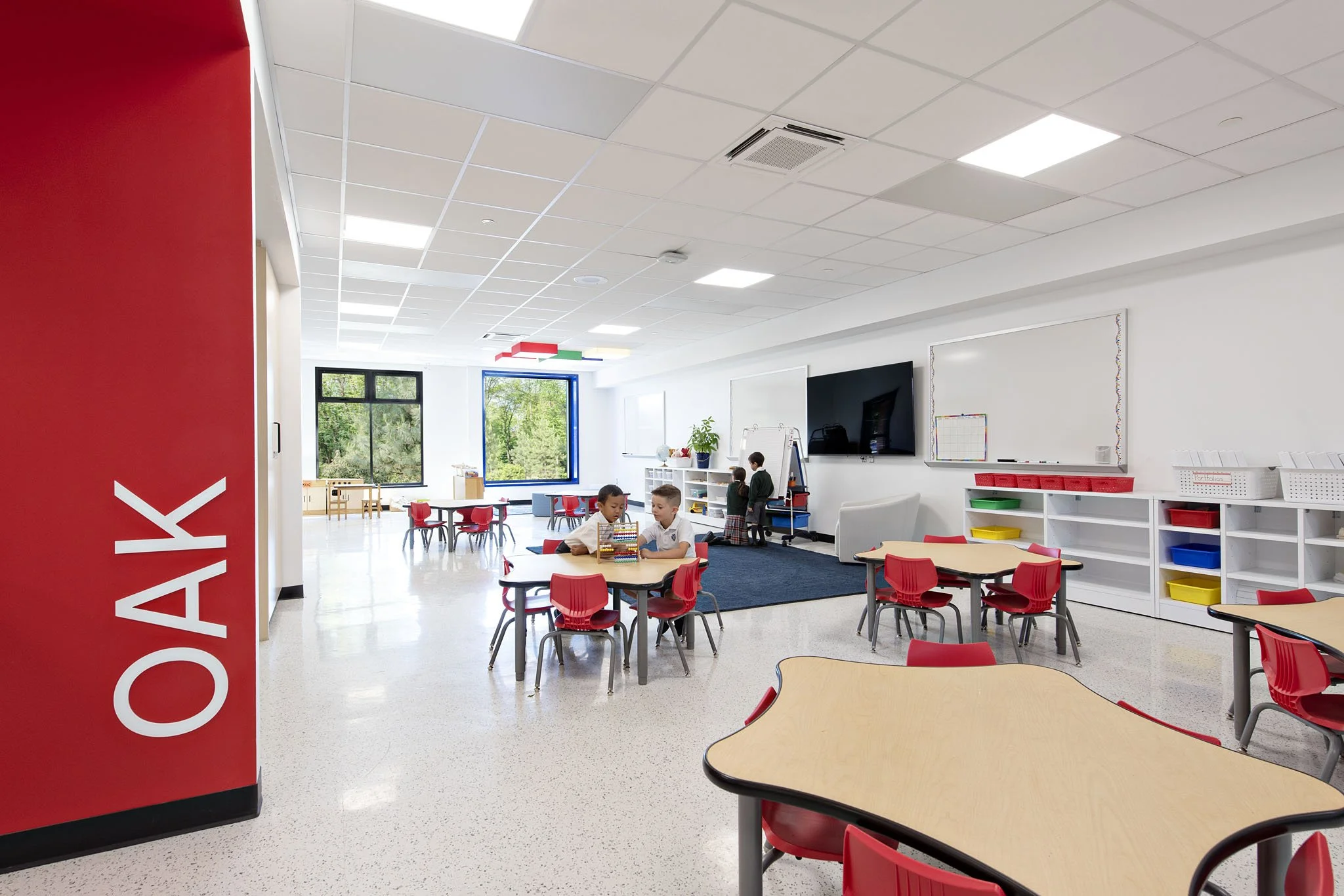 Interior of a modern classroom with students at tables, a large TV, whiteboards, and colorful storage bins, with large windows showing greenery outside.