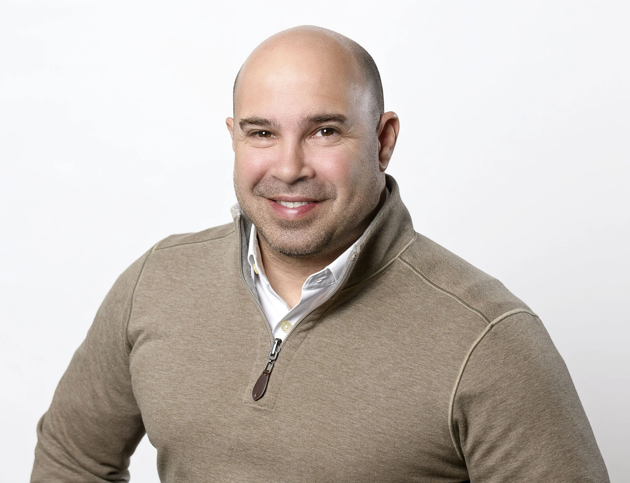 A smiling man with a shaved head, wearing a beige zip-up sweater over a white collared shirt, in front of a plain white background.
