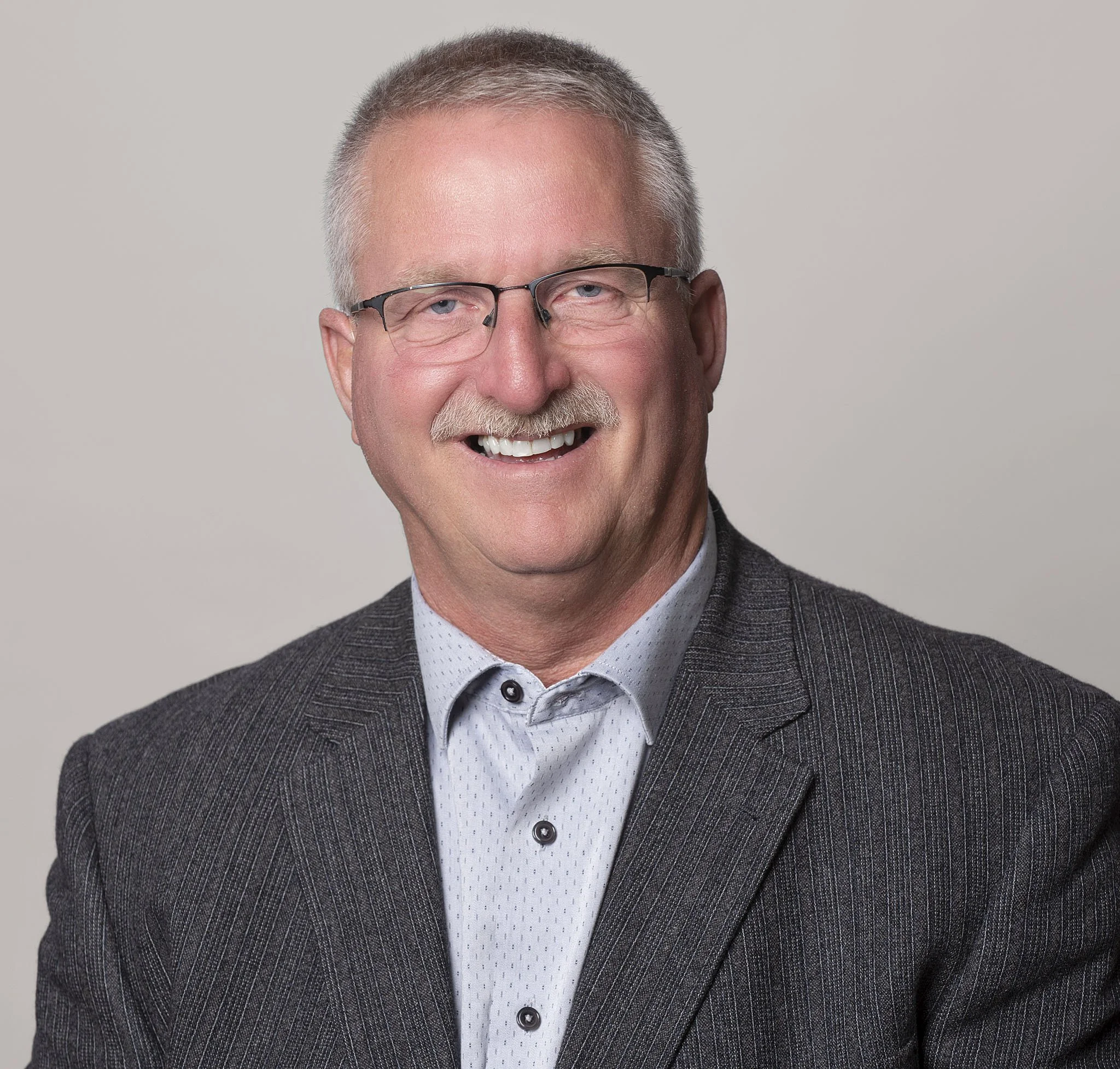 Close-up of a smiling older man with gray hair, glasses, and a gray suit jacket.