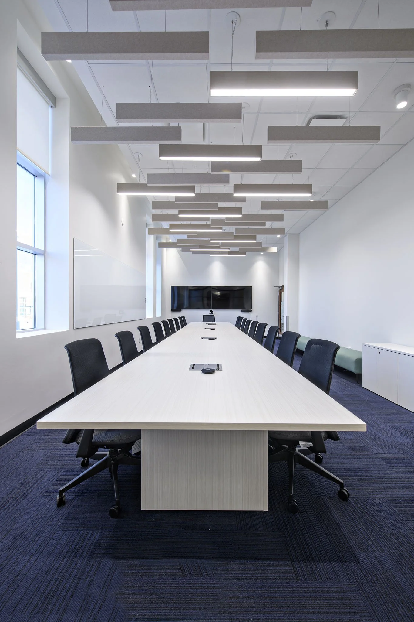 A modern conference room with a long white table, black chairs, large windows, and suspended rectangular light fixtures.