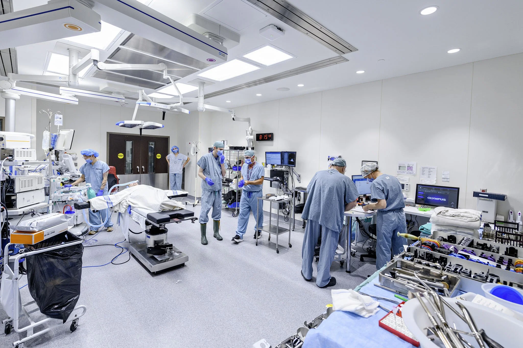 Image of a modern operating room with medical staff performing surgery. The room is equipped with surgical lights, monitors, and various medical equipment, with several surgeons and nurses attending to a patient.