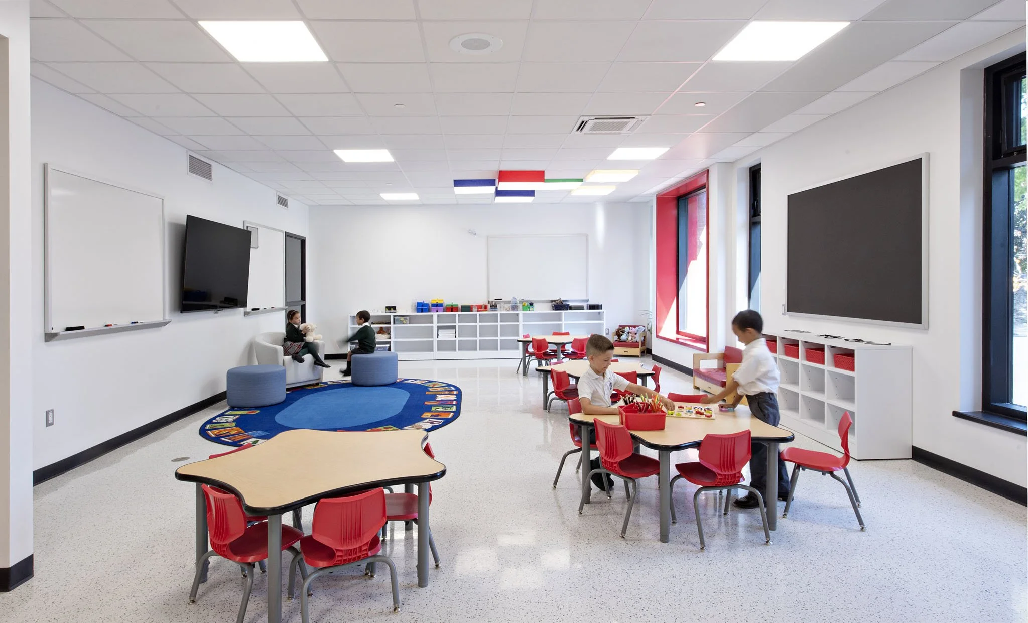 Children playing and engaging in activities in a brightly lit classroom with white walls, large windows, and colorful decor, including a blue carpet with a rainbow border, red chairs, and white shelving filled with toys and supplies.