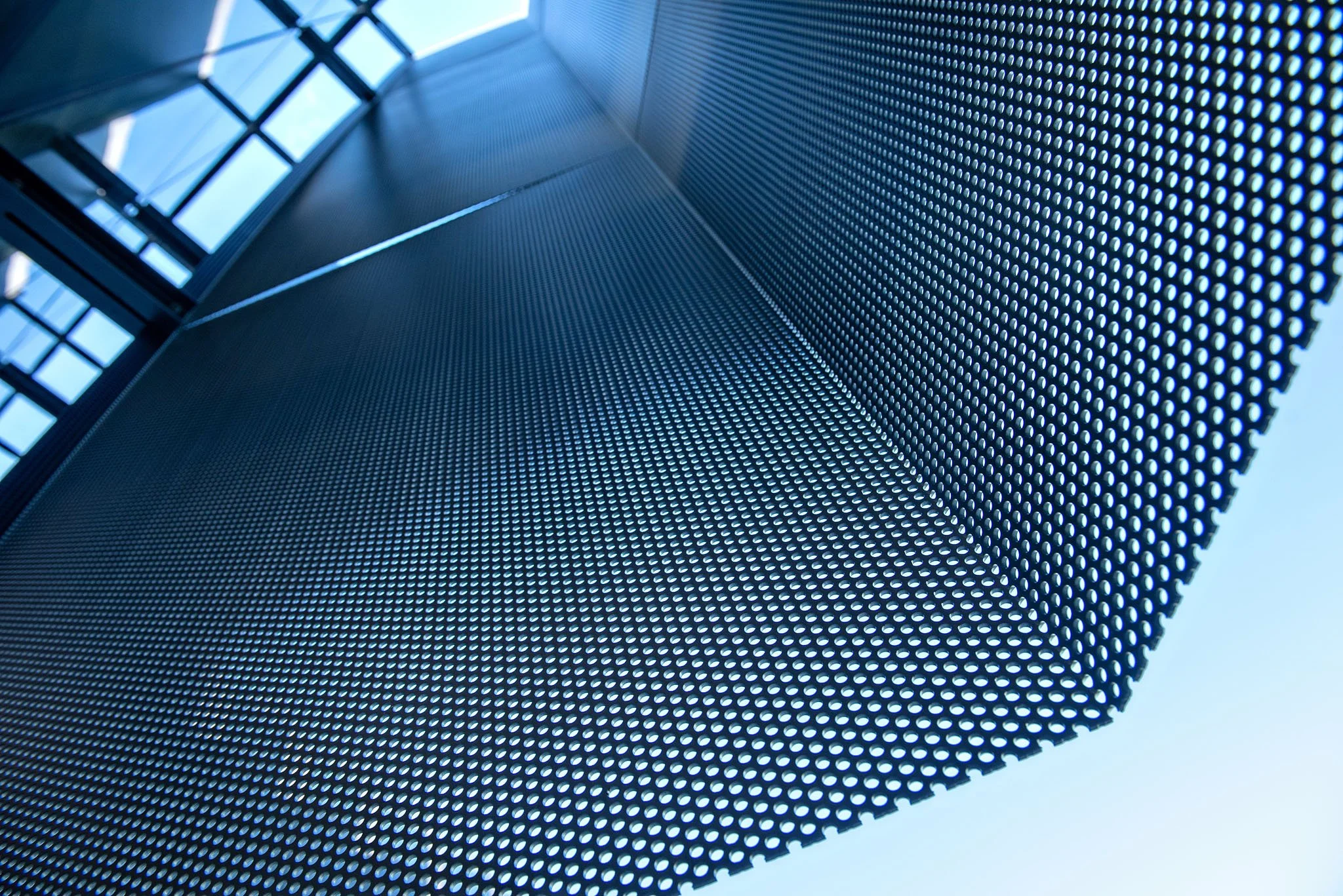Close-up view of a perforated metal surface with circular holes, reflecting sunlight and showing parts of a building structure against a blue sky.