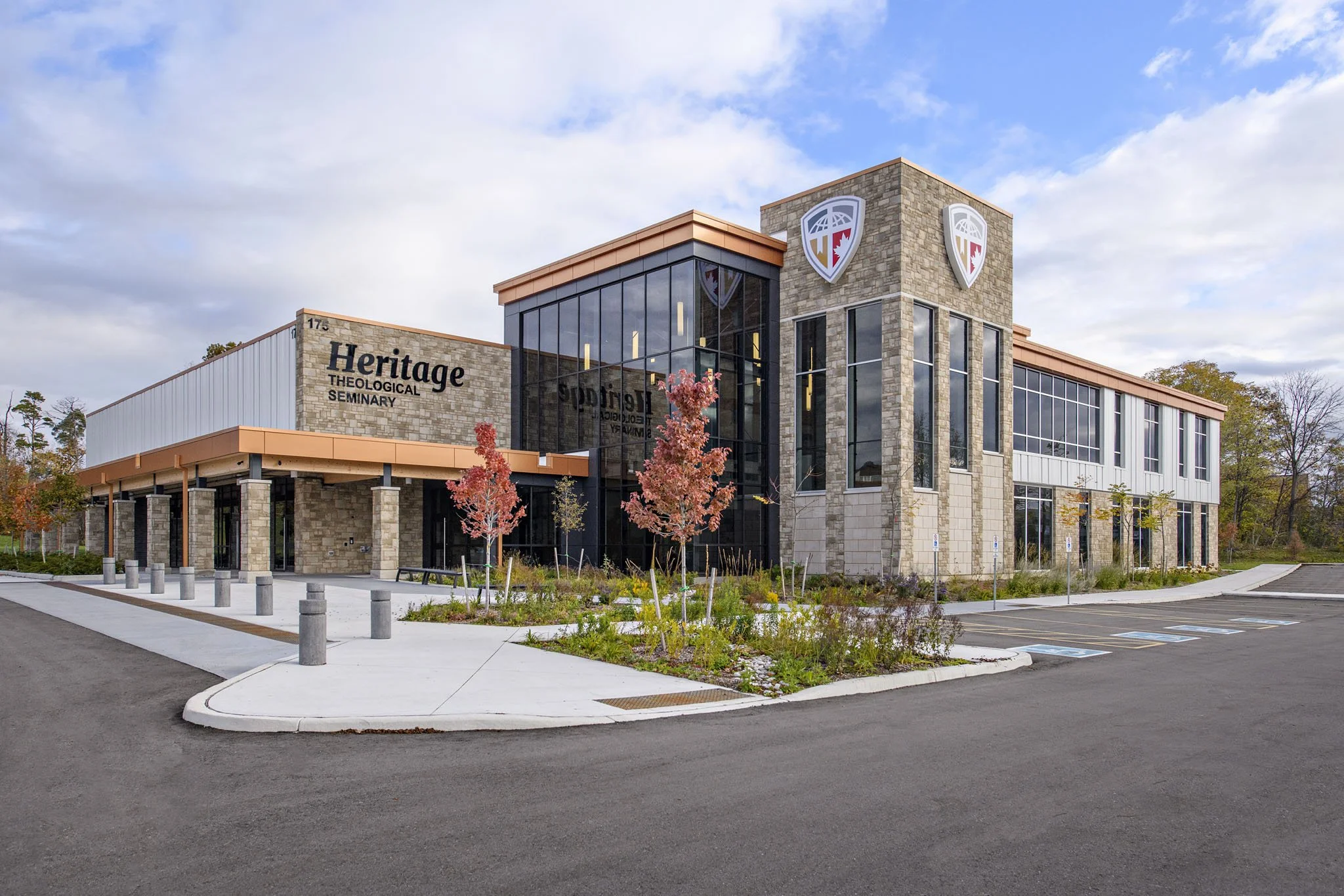 Front view of Heritage Theological Seminary building with parking lot and landscaped area, blue sky with clouds, and autumn trees.
