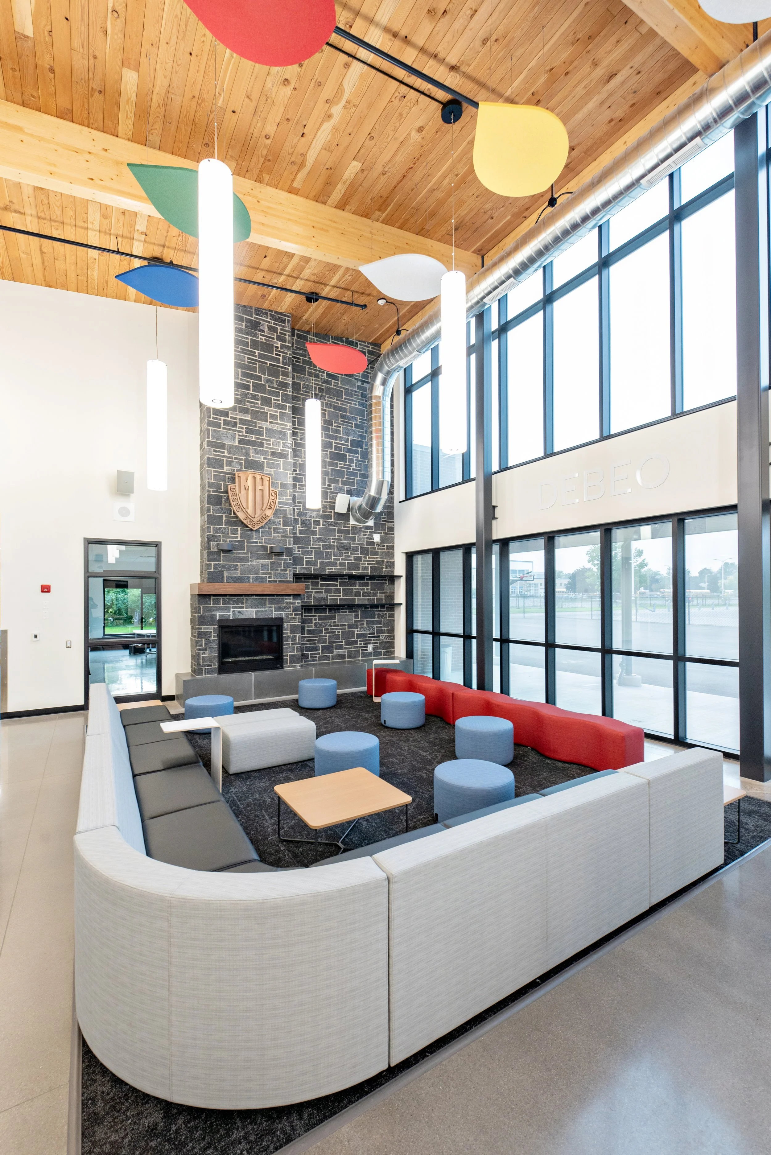 A modern lobby with large windows, a stone fireplace, and colorful seating arranged in a semi-circle. There are pendant lights hanging from a wooden ceiling with colorful abstract shapes. The lobby has a mix of contemporary furniture and a shield-sha