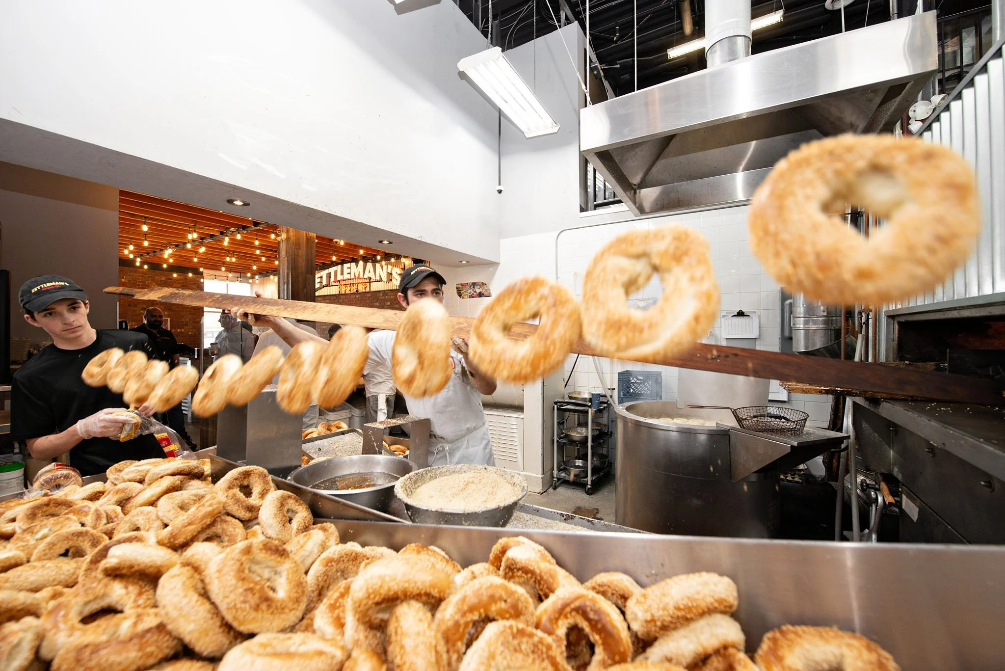 Workers preparing bagels in a commercial kitchen, with a background sign reading 'Tittleman's' and string lights. Commercial photography 