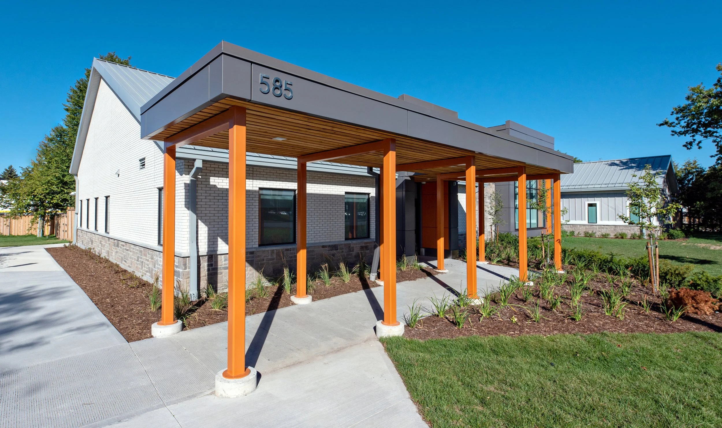 Modern building with white brick walls, gray roof, and orange support columns at the entrance, surrounded by a landscaped yard with young plants and trees, under a clear blue sky.