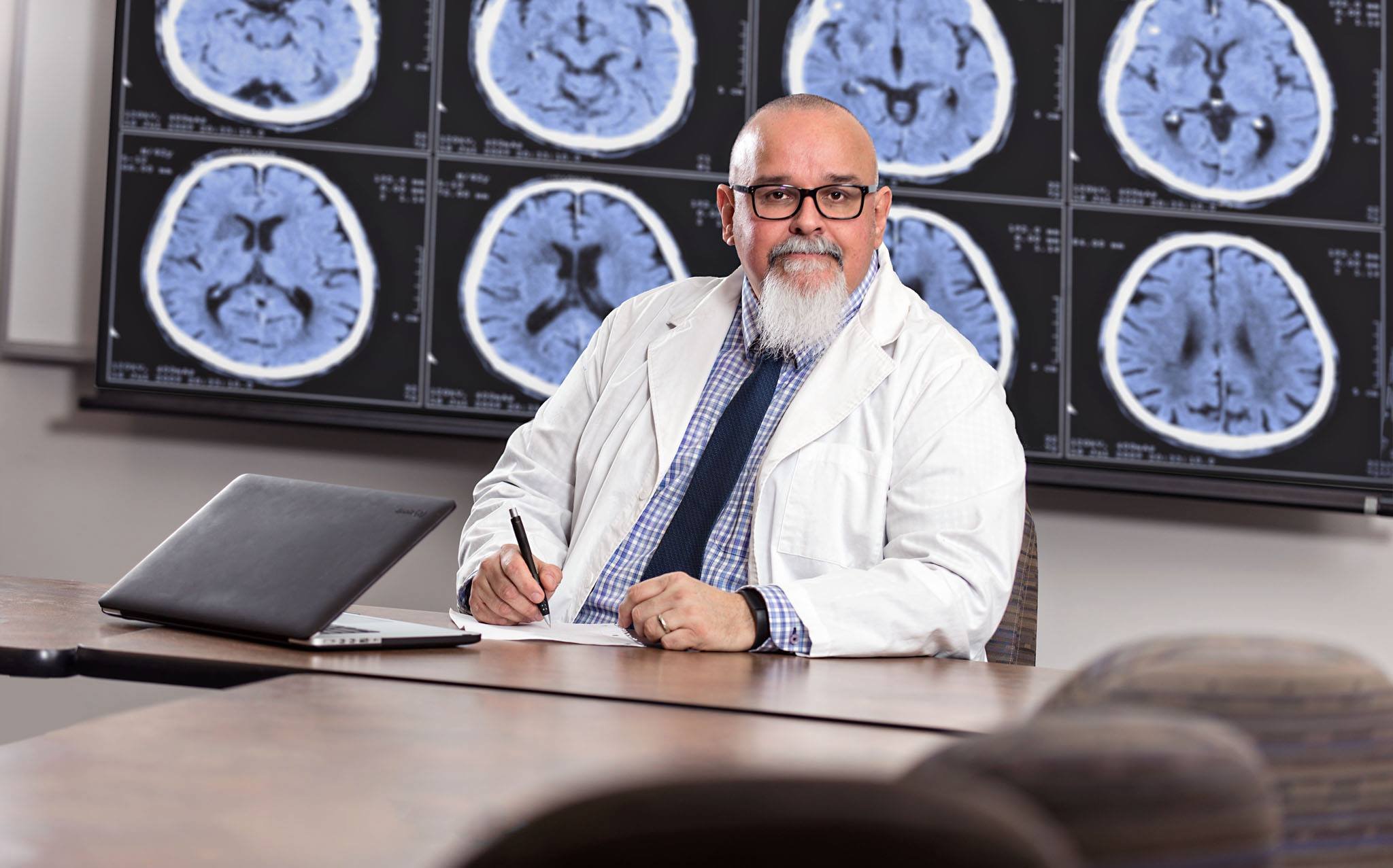 A doctor, white beard is sitting at a desk in front of a large screen displaying brain scans. He is wearing a white lab coat, a checkered shirt, and a dark tie, holding a pen, with a laptop and papers on the desk. Executive portrait photo
