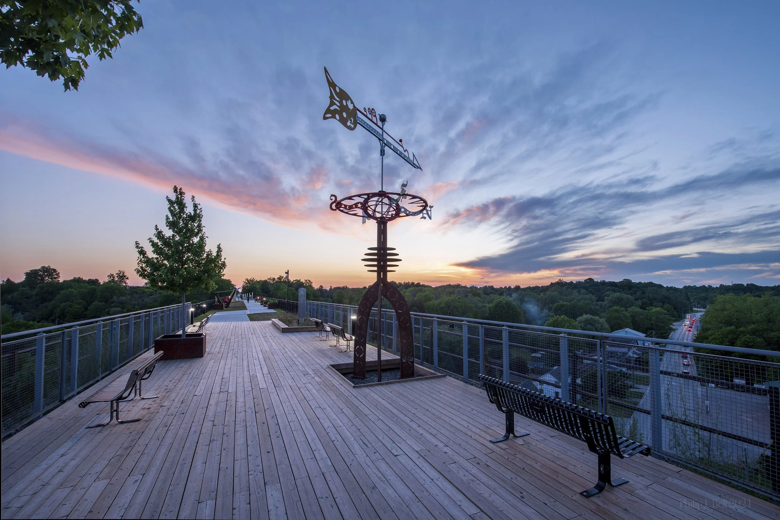 Footbridge overlooking a scenic landscape at sunset with benches along the walkway, a meteorological vane, and a view of trees, hills, and a road with cars.