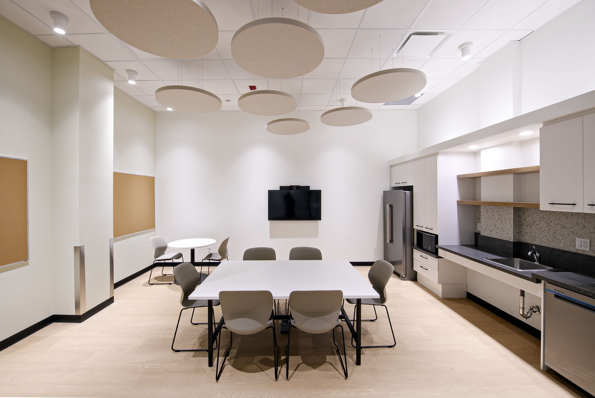 Modern kitchen and dining area with white tables and chairs, beige acoustic ceiling panels, white cabinets, stainless steel refrigerator, TV on the wall, and a kitchenette with a sink and microwave.