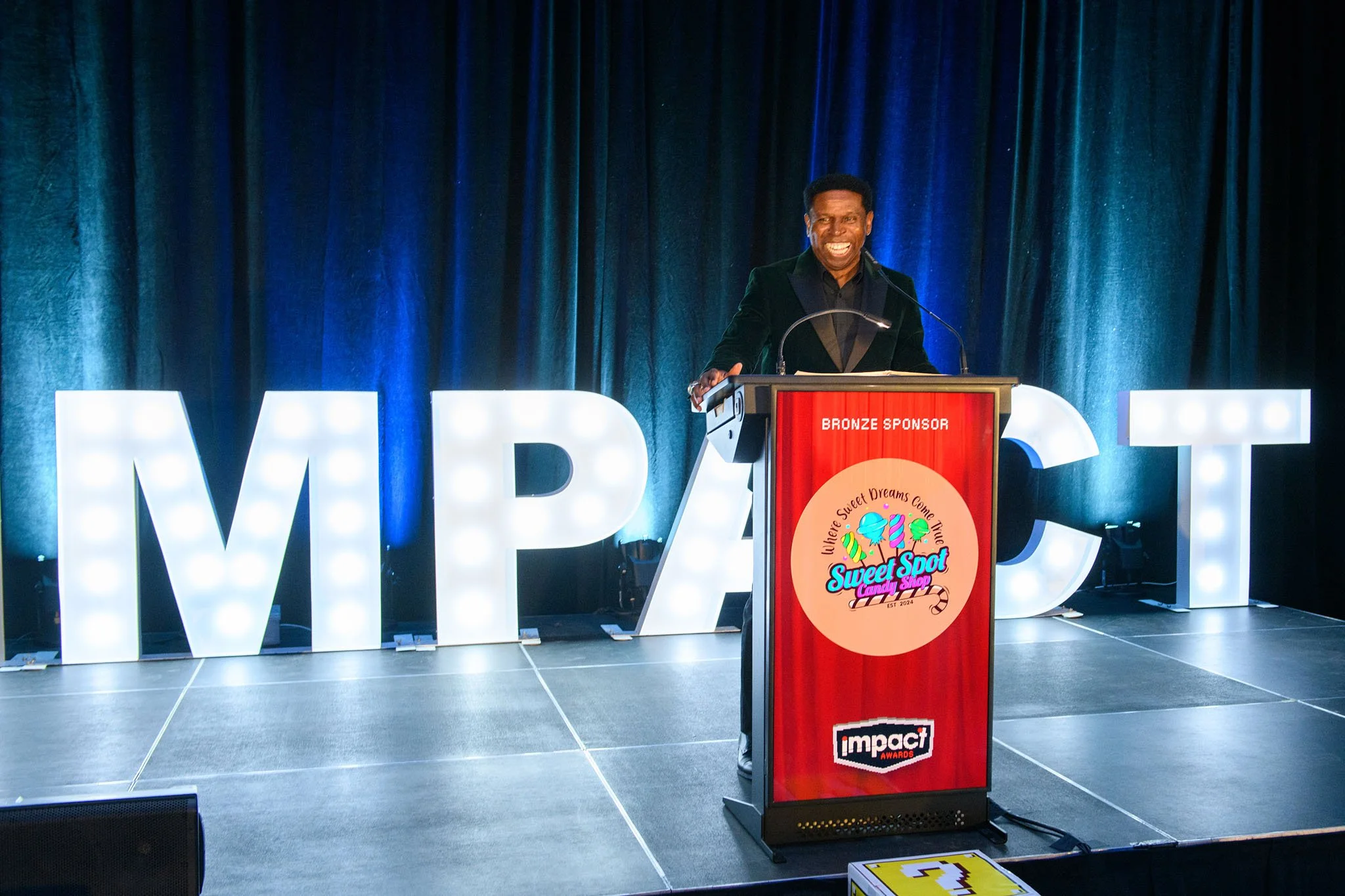 Man smiling at a podium at an awards event with large decorative letters in the background and candy shop logo on the podium.