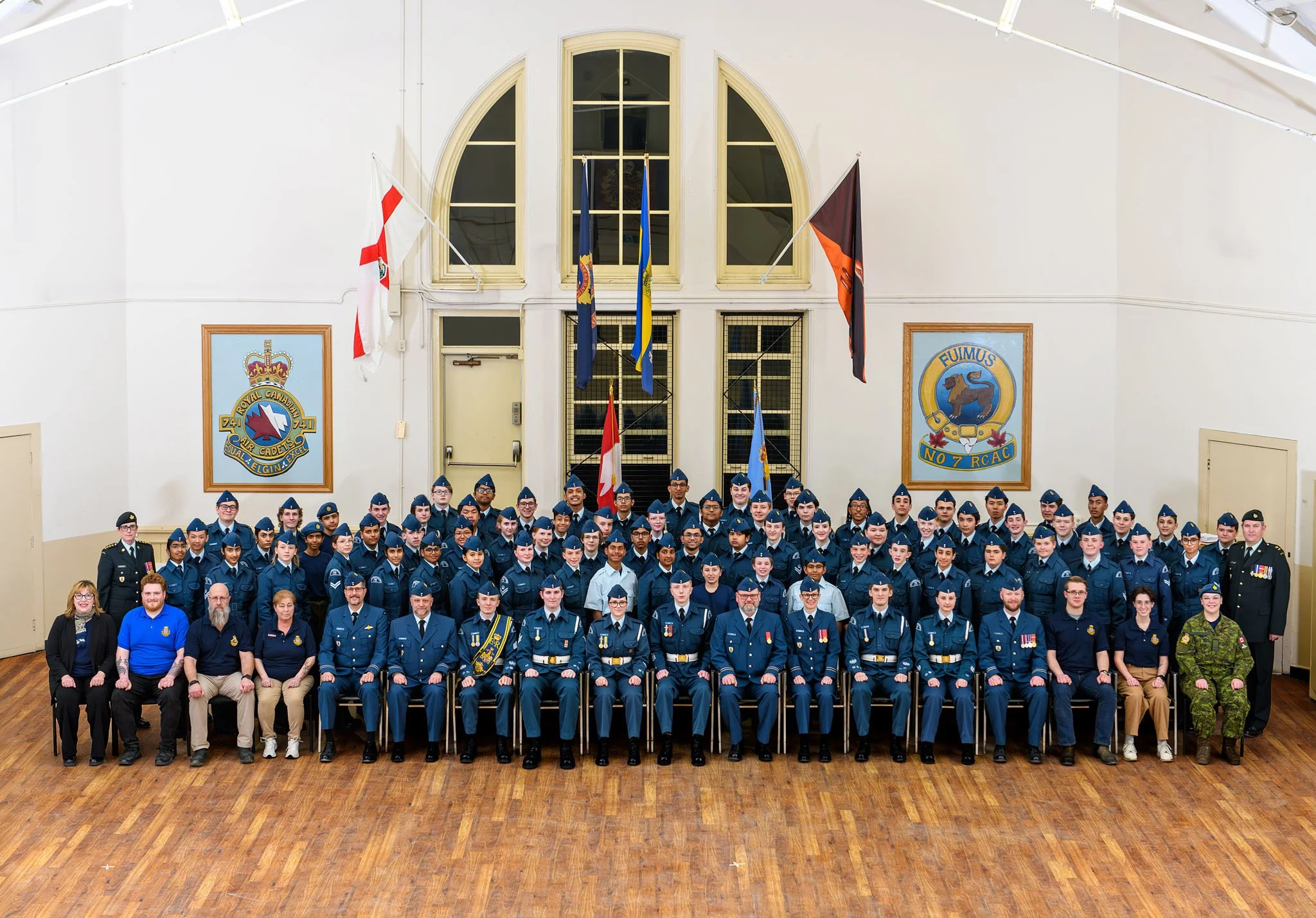 Group photo of cadets in uniform at a ceremony inside a hall with flags and banners, some adults seated in front.