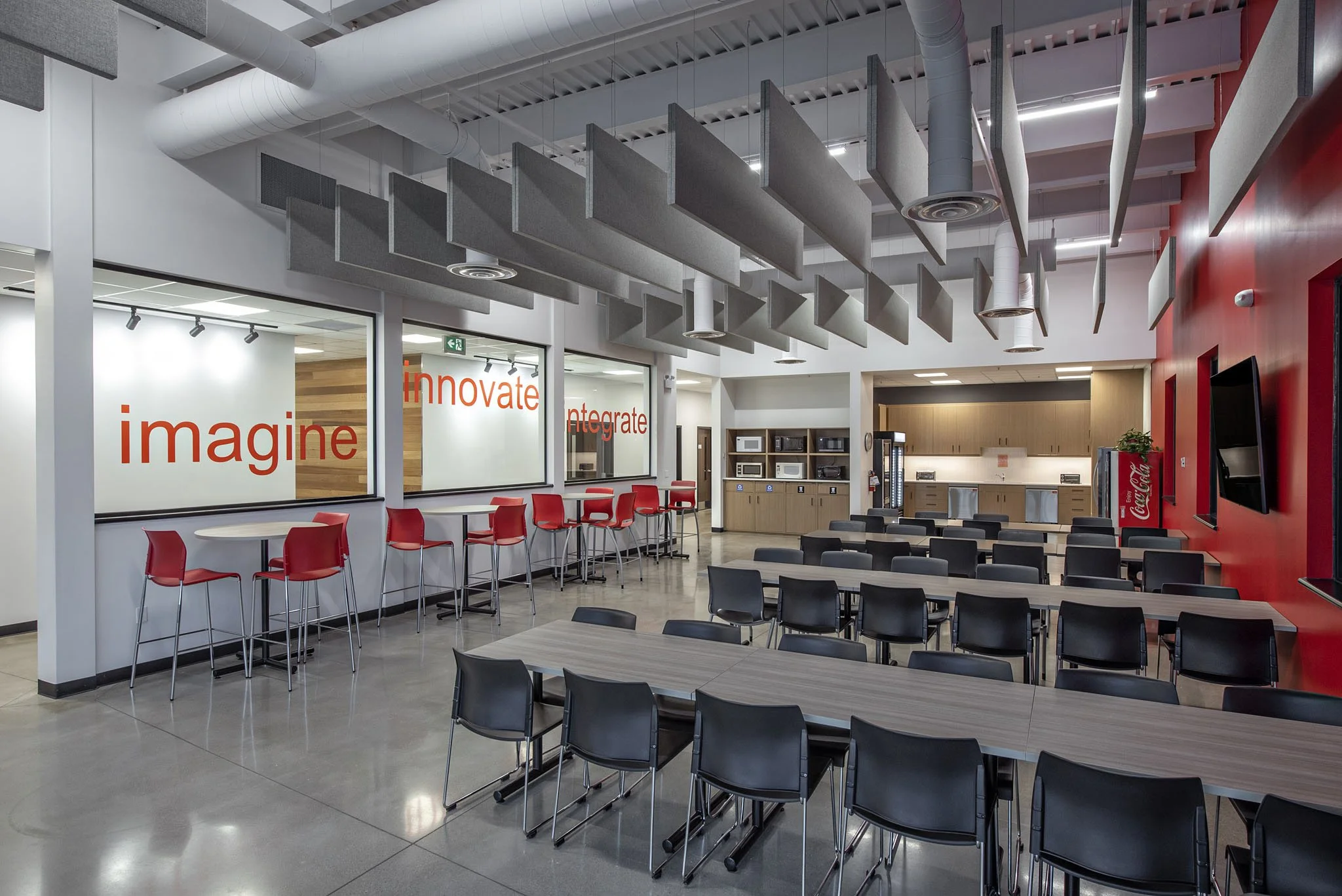 A modern cafeteria with black and red chairs, tables, a red wall with a Coca-Cola vending machine, and a kitchen area with wooden cabinets and microwaves. Ceiling has grey acoustic panels and exposed ducts. Architectural photography. 