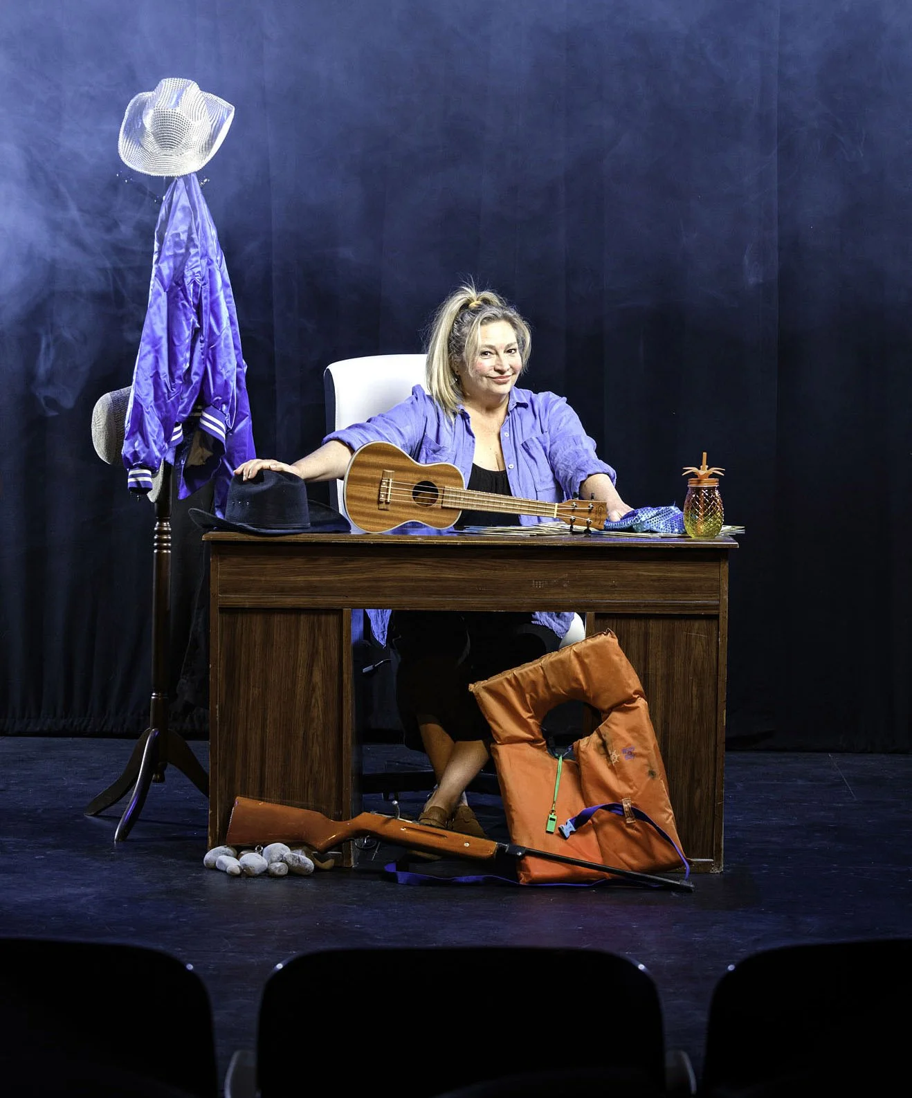 A woman sitting behind a desk with a ukulele, wearing a blue shirt and smiling. Items on the desk include a black hat, a pineapple-shaped container, and a blue cloth. Behind her is a coat rack with a blue jacket and a hat. On the floor under the desk
