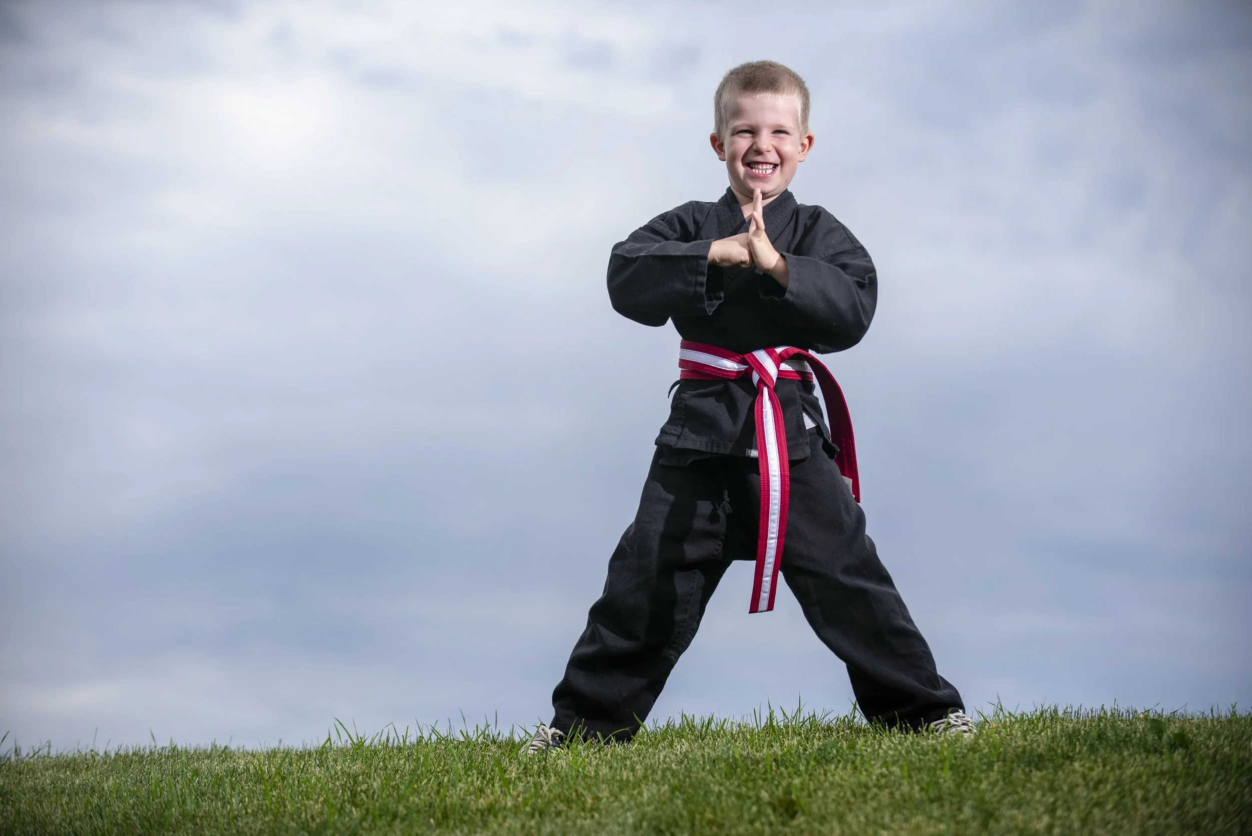 A young boy in a martial arts uniform with a red belt practicing karate outdoors on grass, smiling with his hands in a prayer position.