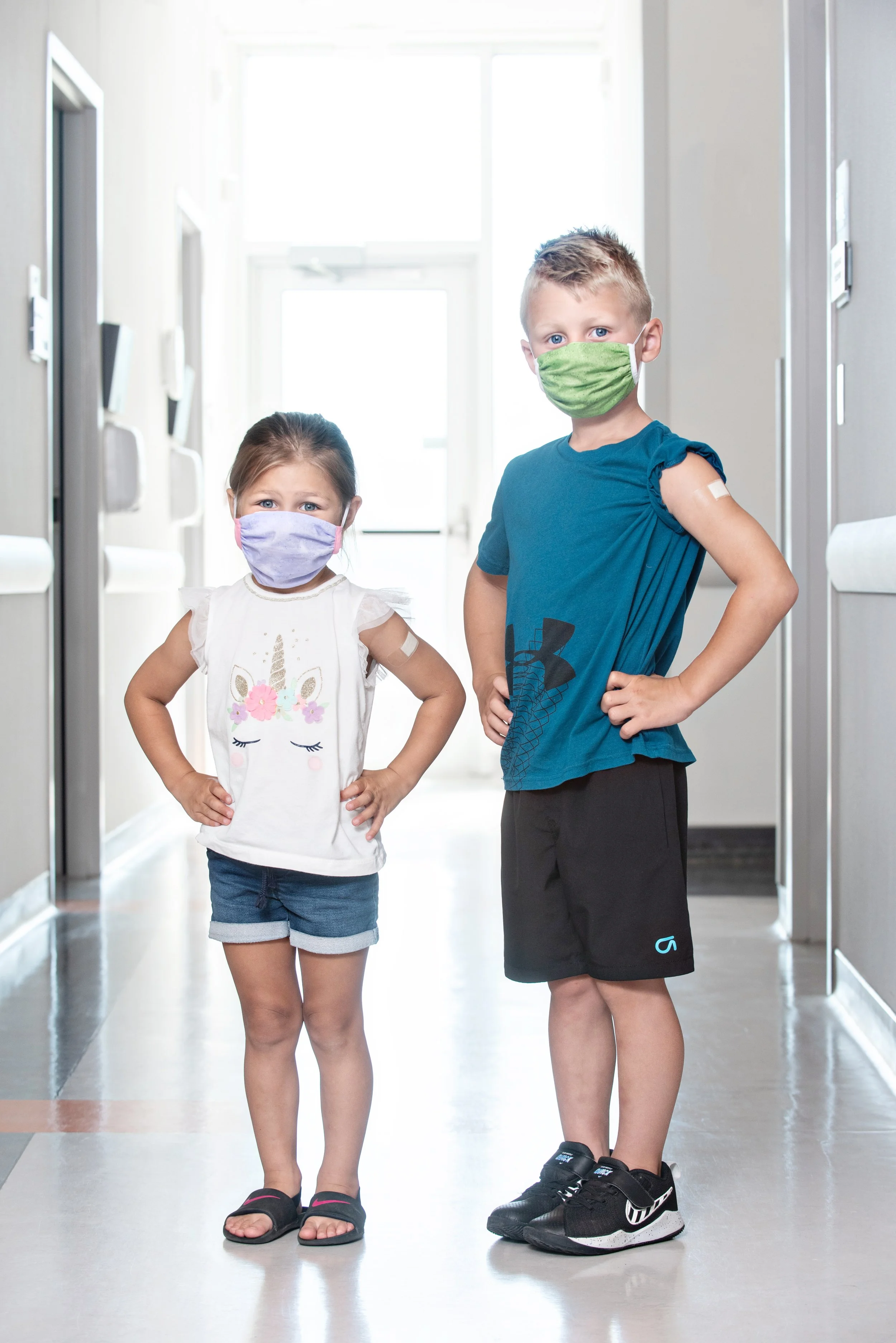 Two children, boy and girl, standing in a hallway, wearing face masks, with the boy having a bandage on his upper arm, and both with their hands on their hips.