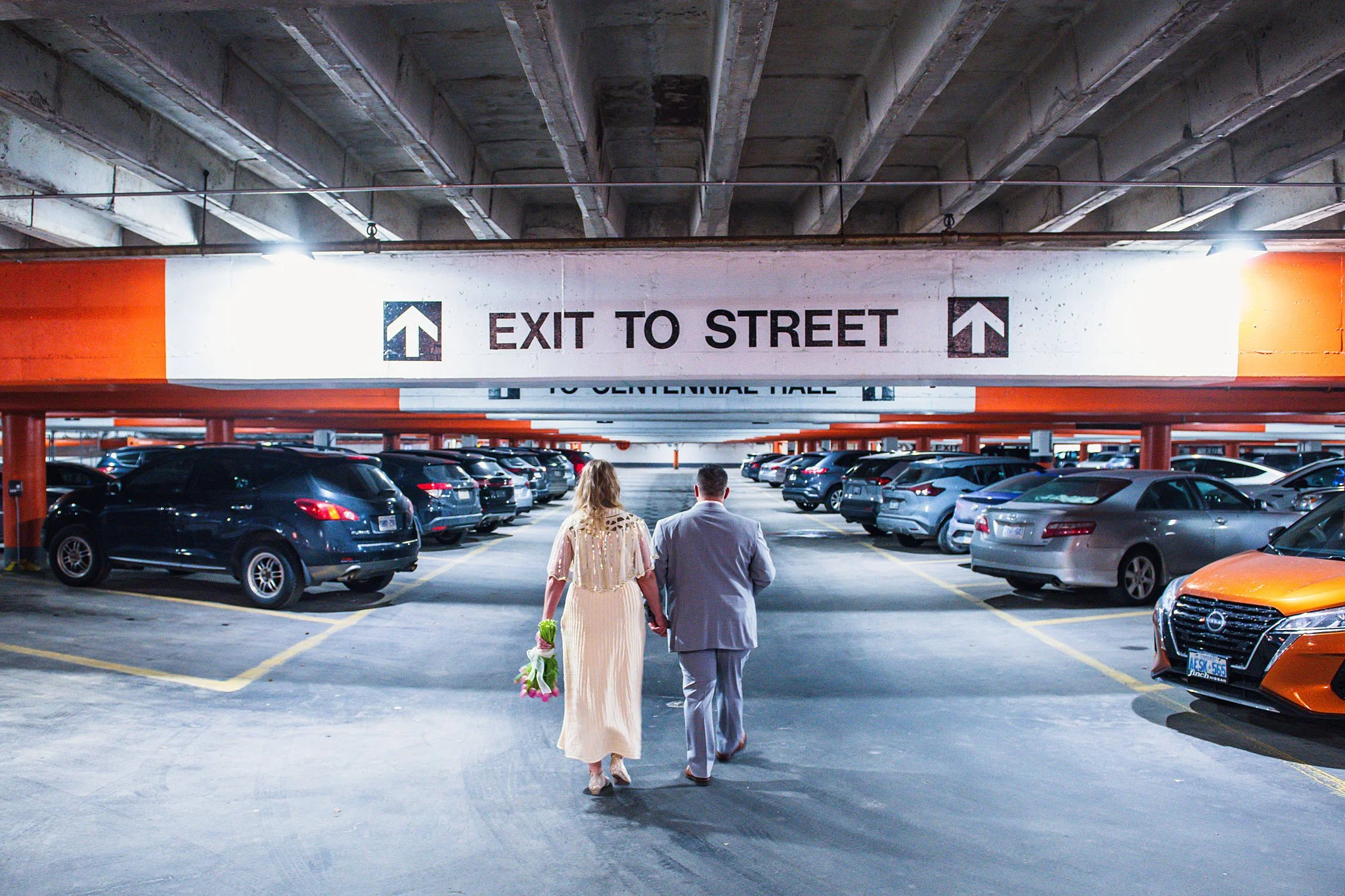 A couple dressed as a bride and groom holding hands and walking through a parking garage area with many parked cars, heading towards an exit.