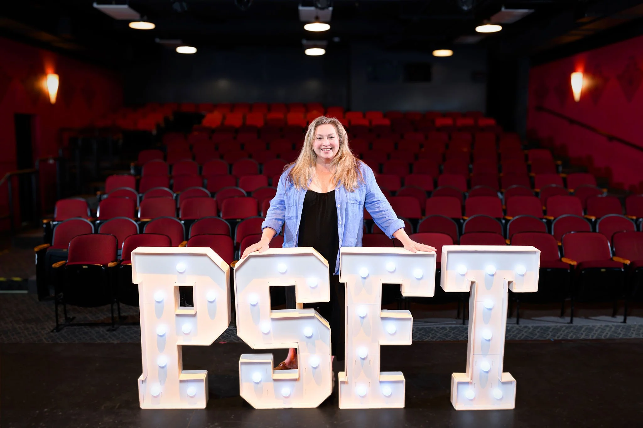 A woman with long blonde hair smiling and standing behind large illuminated letters spelling 'LOVE' in a theater with red seats and dark walls.