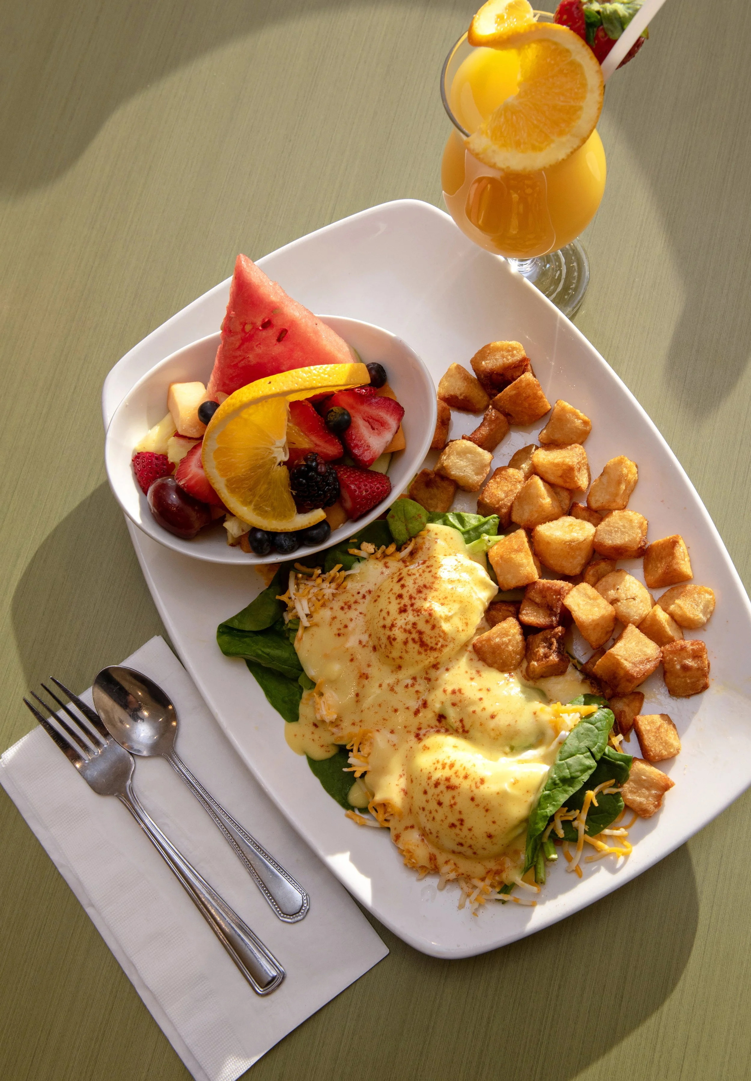 A breakfast plate with eggs Benedict, crispy hash browns, a side salad, and a bowl of fresh fruit with watermelon, strawberries, blueberries, blackberries, and orange slices, accompanied by a tall glass of orange juice garnished with orange slices an
