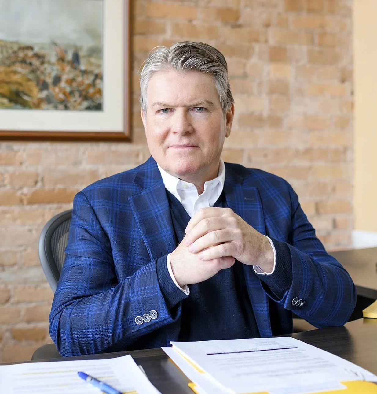A middle-aged man in a blue plaid blazer and white shirt sitting at a desk with documents and a pen, clasping his hands together, in front of a brick wall. A framed landscape painting hangs on the wall behind him.