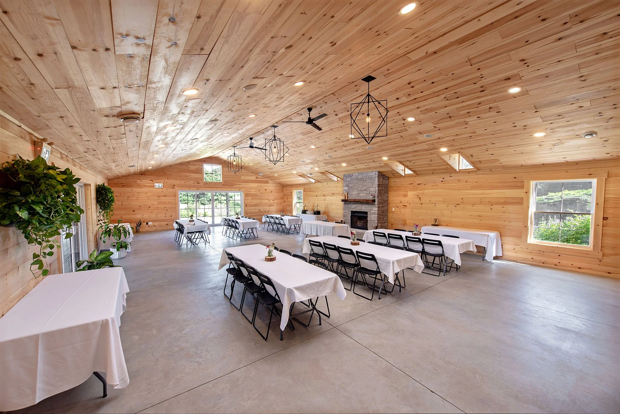 Interior of a banquet hall with wooden ceiling and walls, decorated with hanging geometric light fixtures, ceiling fans, and windows, set up with tables covered with white tablecloths and black chairs.