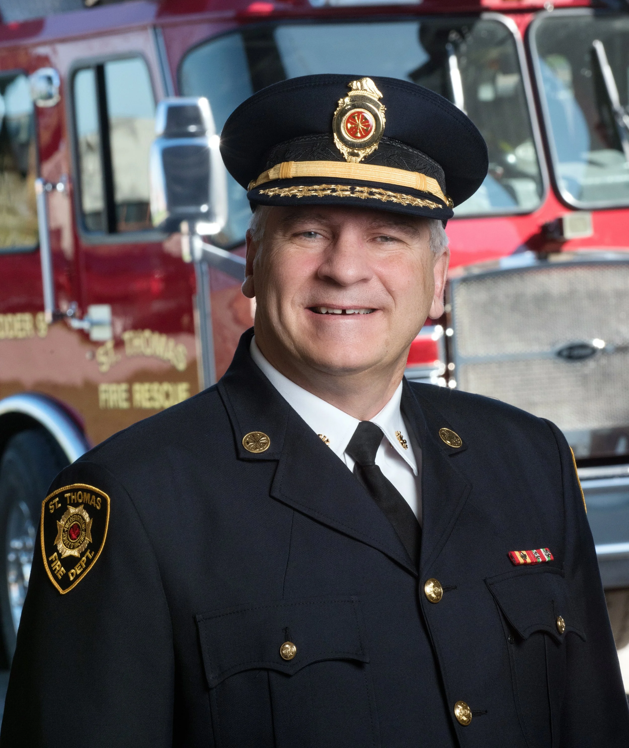A firefighter in formal dress uniform, including a blue hat with badge, standing in front of a red fire truck. Executive portrait photography. 