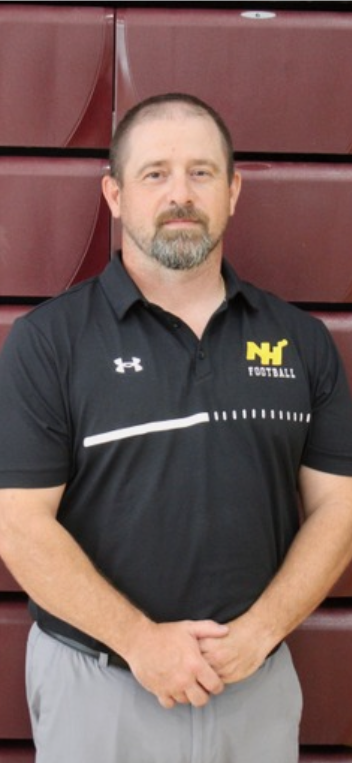A man standing indoors in front of maroon-colored bleachers, wearing a black football coach polo shirt with a logo on the chest, gray pants, and with short brown hair and a beard.