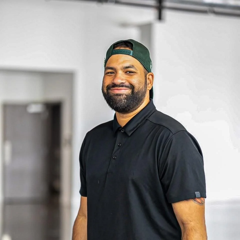 A smiling man wearing a black polo shirt and a green baseball cap backward in a modern indoor setting.