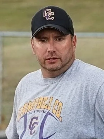 A man wearing a black baseball cap with the letters 'EC' and a gray t-shirt with yellow and purple lettering standing outdoors in a grassy area.