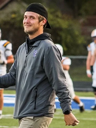 A smiling man wearing a black cap, gray jacket, and khaki pants standing on a sports field with football players in the background.