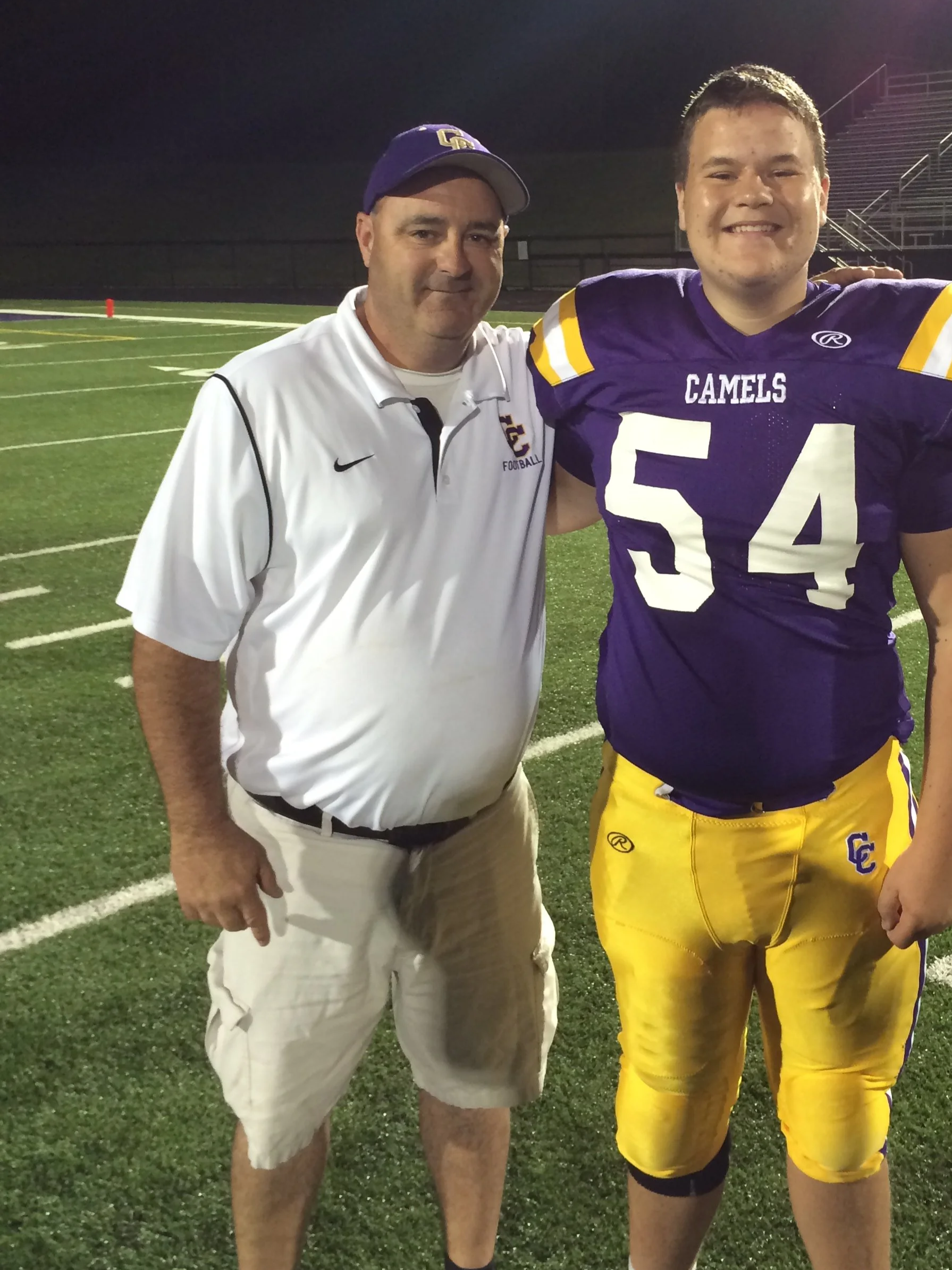 Two men standing on a football field at night, smiling for the camera. One is wearing a white polo shirt and khaki shorts, and the other is in a purple and yellow football uniform with the number 54. The background shows football field markings and empty bleachers.