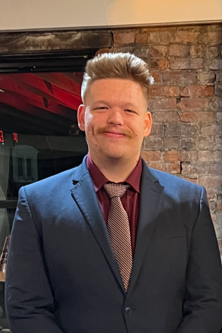 Young man with styled hair and a mustache wearing a navy blazer, maroon shirt, and tie, standing indoors in front of a brick wall.