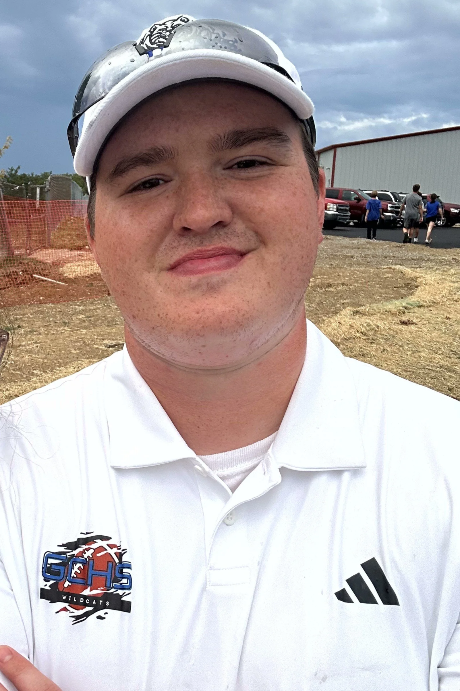 A close-up of a young man wearing a white sports polo shirt with a logo that says "GHS Wildcats" and a black Adidas logo, a white baseball cap, and sunglasses on top of the cap. He is outdoors near a parking lot with other people and vehicles in the background, with cloudy skies overhead.