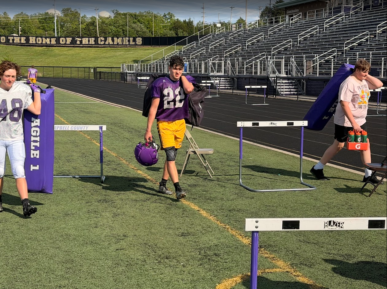 Young football players leaving the field after a game at a stadium, carrying their helmets and water bottles on a sunny day.