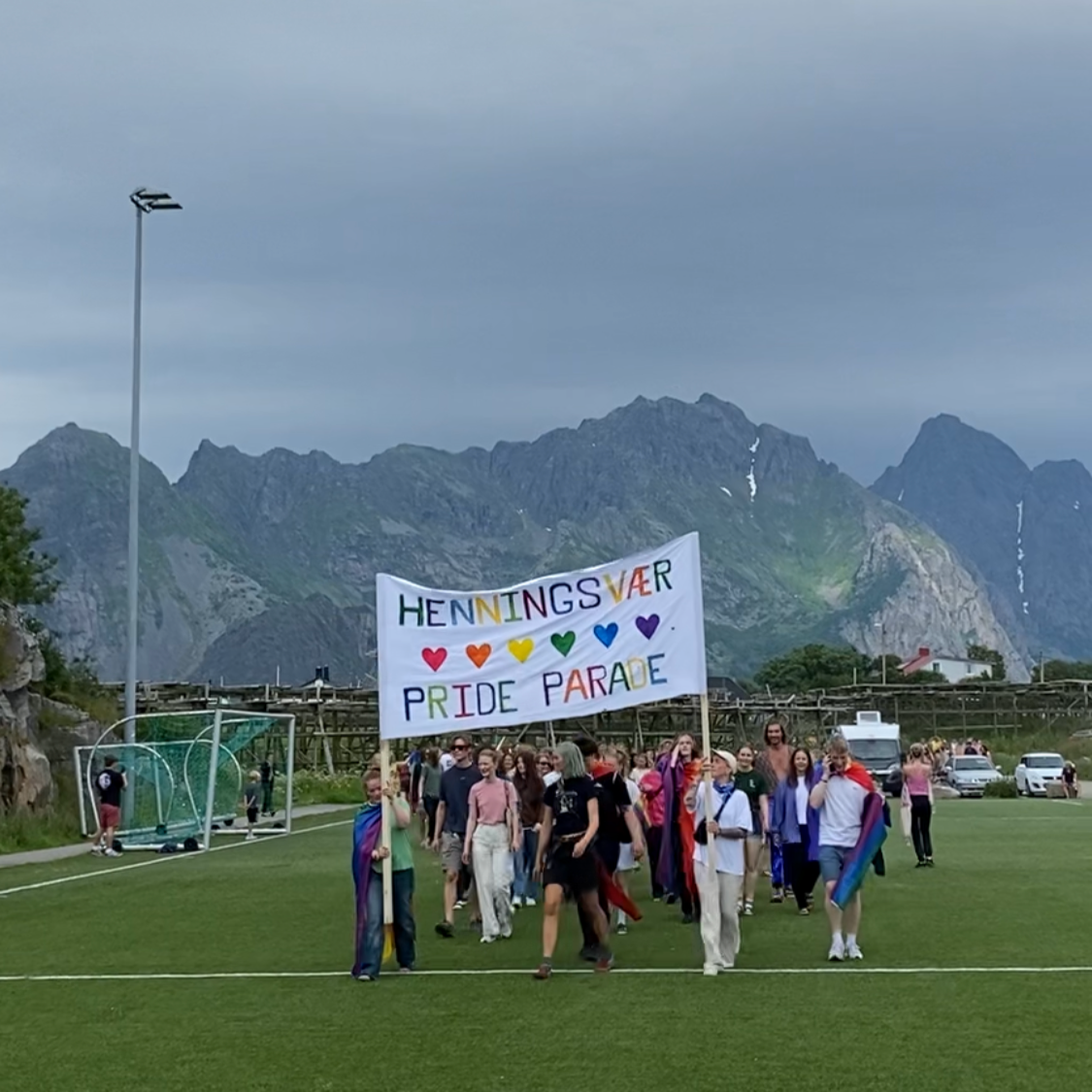 En gruppe mennesker går i en parade på en fotballbane med et banner som sier «HENNINGSVÆR PRIDE PARADE» foran fjell i bakgrunnen, flere av deltakerne har på regnbuearmbånd og flagg.