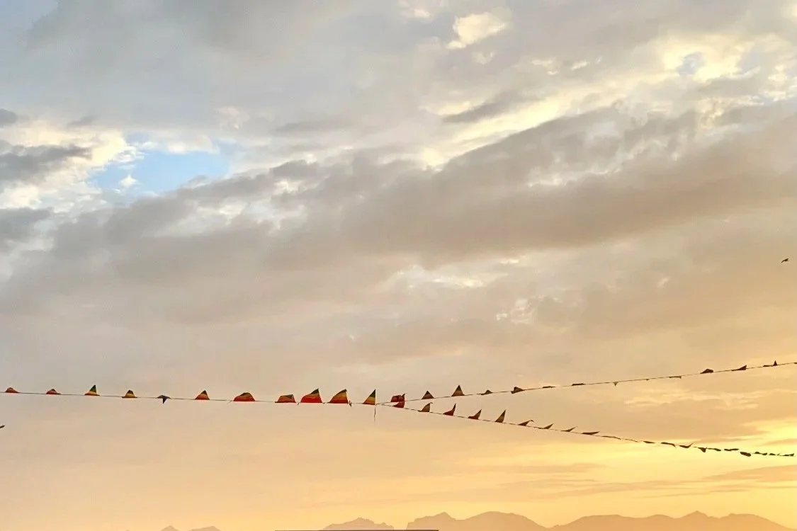 Fargerike flagg hengende i en rekke foran en solnedgang med fjell i bakgrunnen.