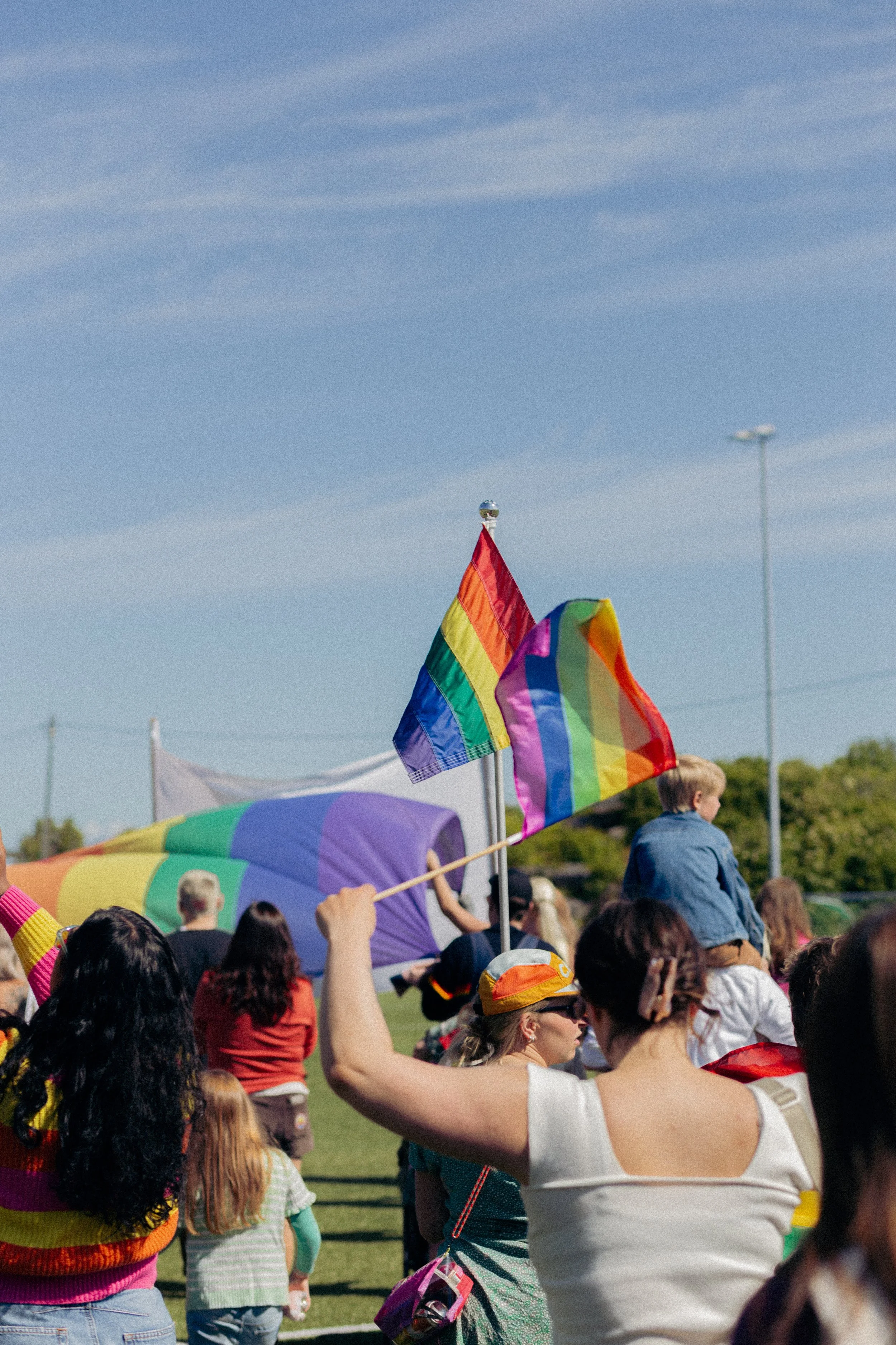  personer på et utendørs arrangement med flere regnbueflagg, folk som ser ut til å delta i en parade eller demonstrasjon, under en klar blå himmel