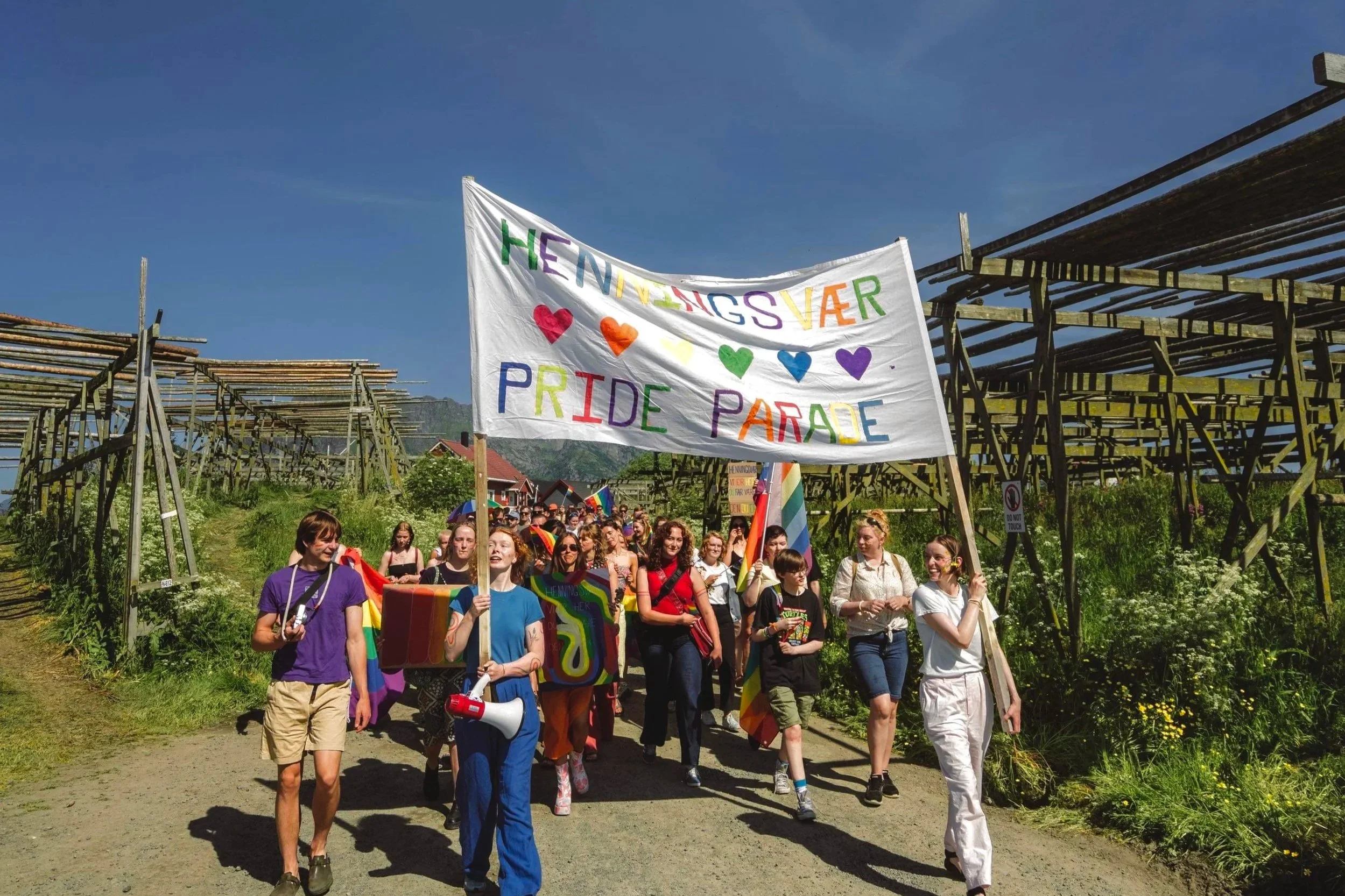 Et Pride-tog med én stor fargerik banner som sier «The Winners Are Pride Parade» blir båret av en gruppe mennesker, mange av dem smiler og viser støtte for LHBTQ+ rettigheter.