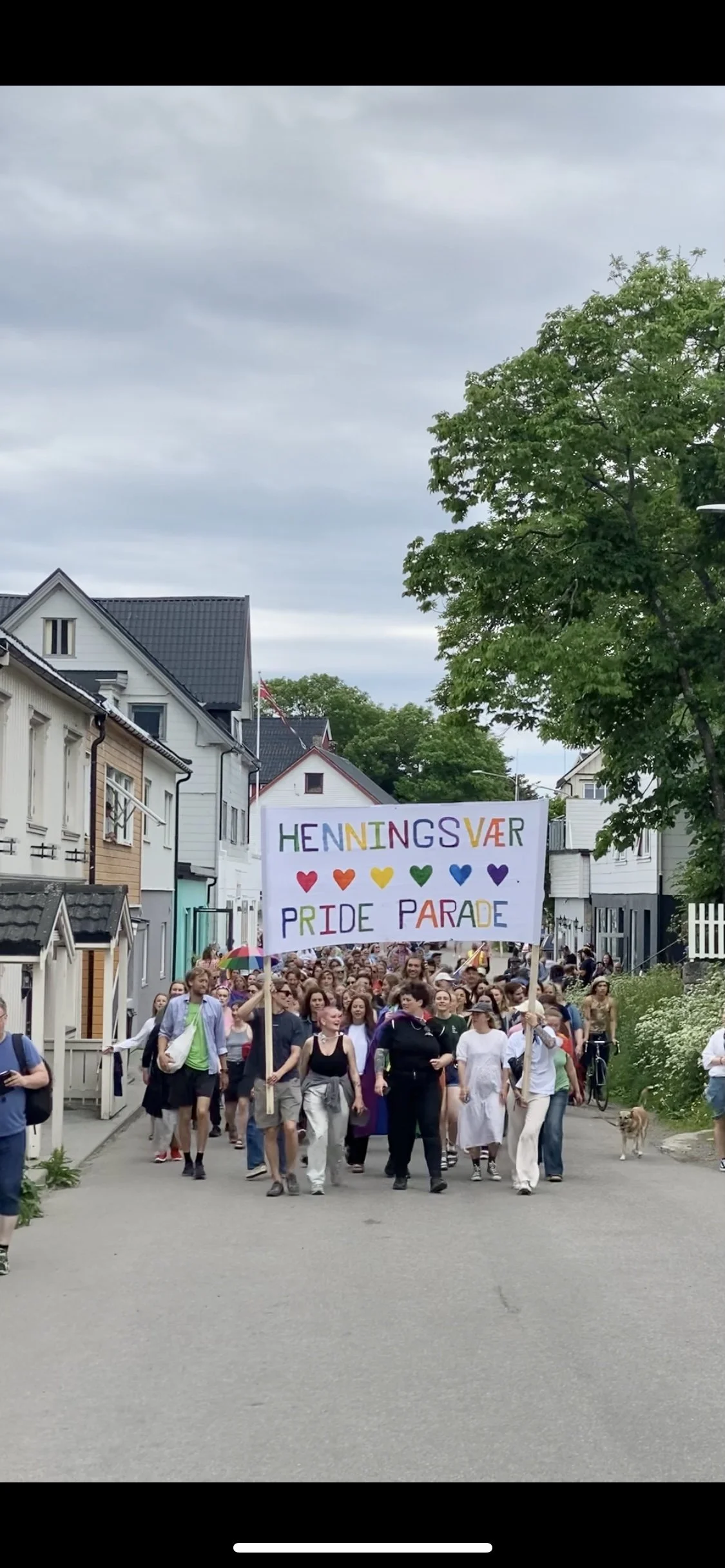 Folk som deltar i Pride-parade, holder et fargerikt skilt med teksten «HENNINGSVÆR PRIDE PARADE» og hjerter i regnbuens farger, på en gate omgitt av boliger.